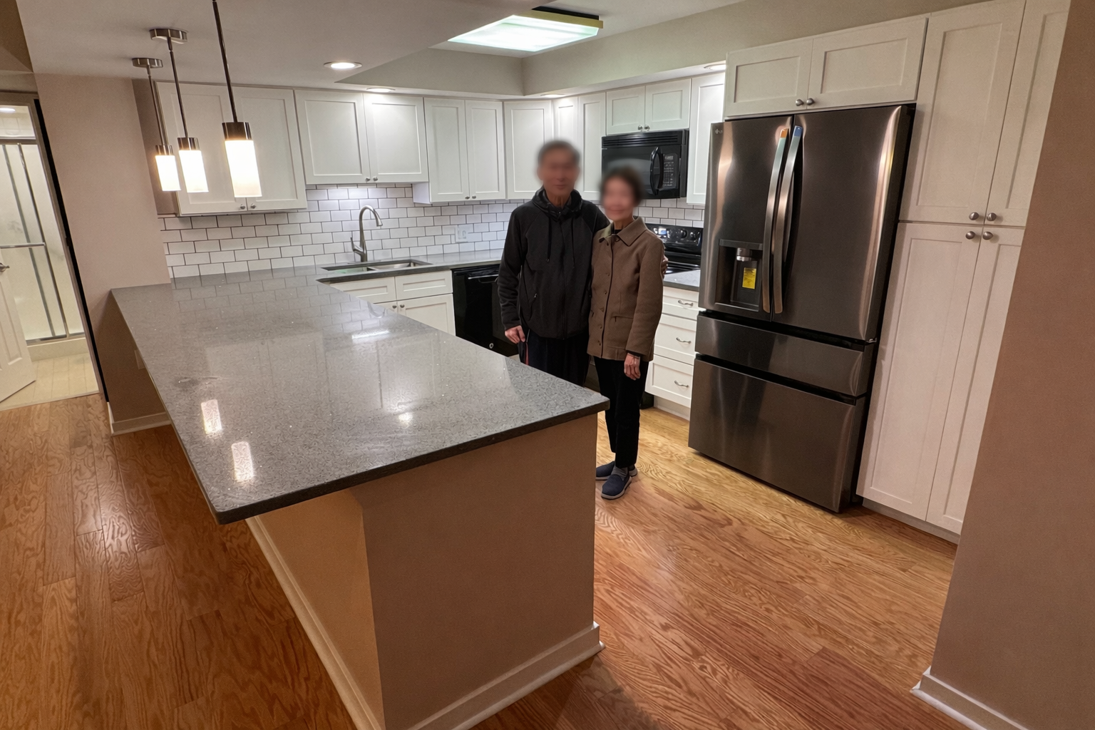 A kitchen with white cabinets, a granite countertop, a stainless steel refrigerator, and wooden flooring. Two people, a man and a woman, are standing together in the kitchen, blurred for privacy. The kitchen has pendant lighting and a white subway ti