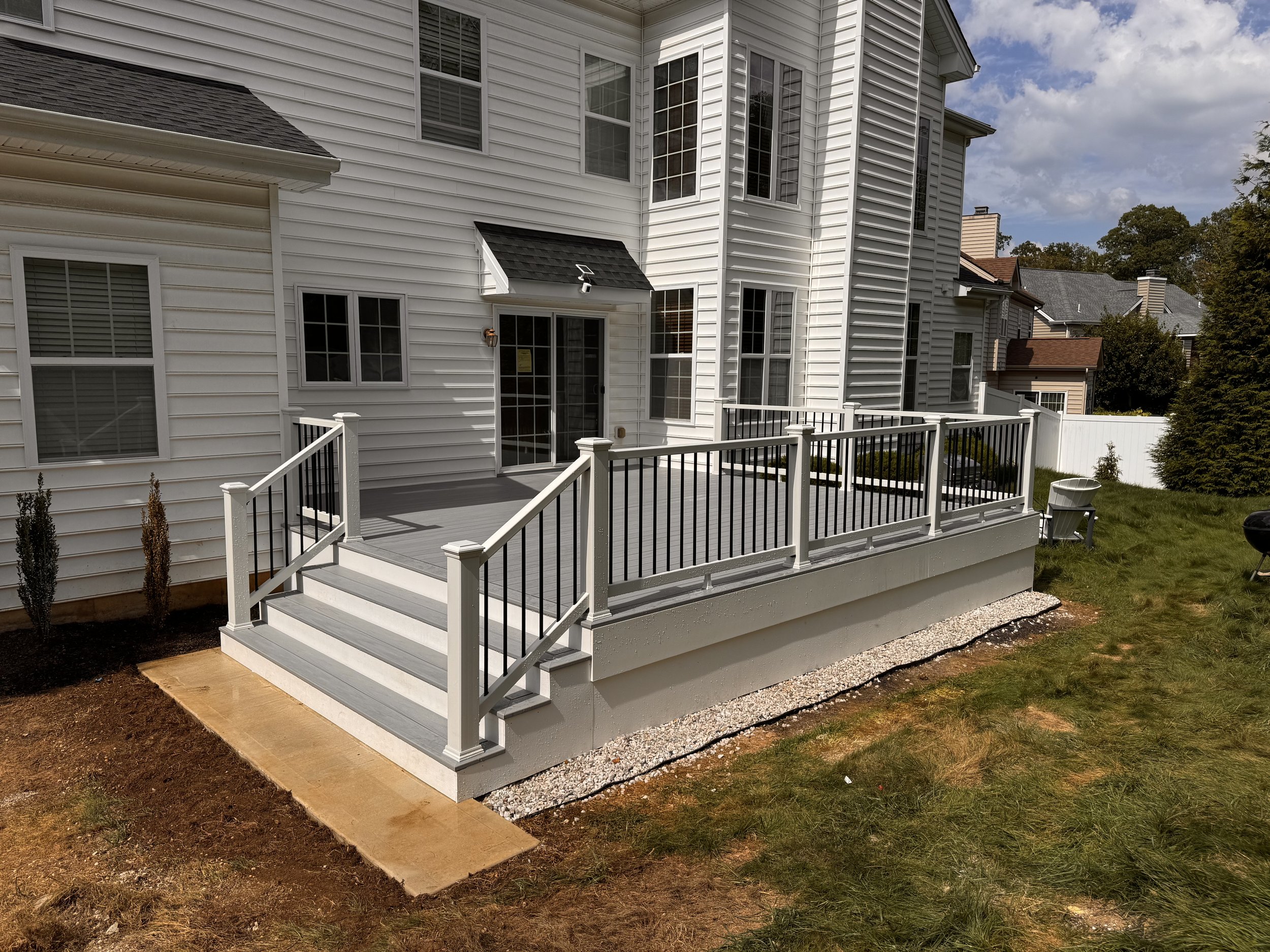 Newly built outdoor deck with white railing and black balusters attached to a white house with large windows, small stairs leading down to a grassy yard, and surrounding neighborhood houses.