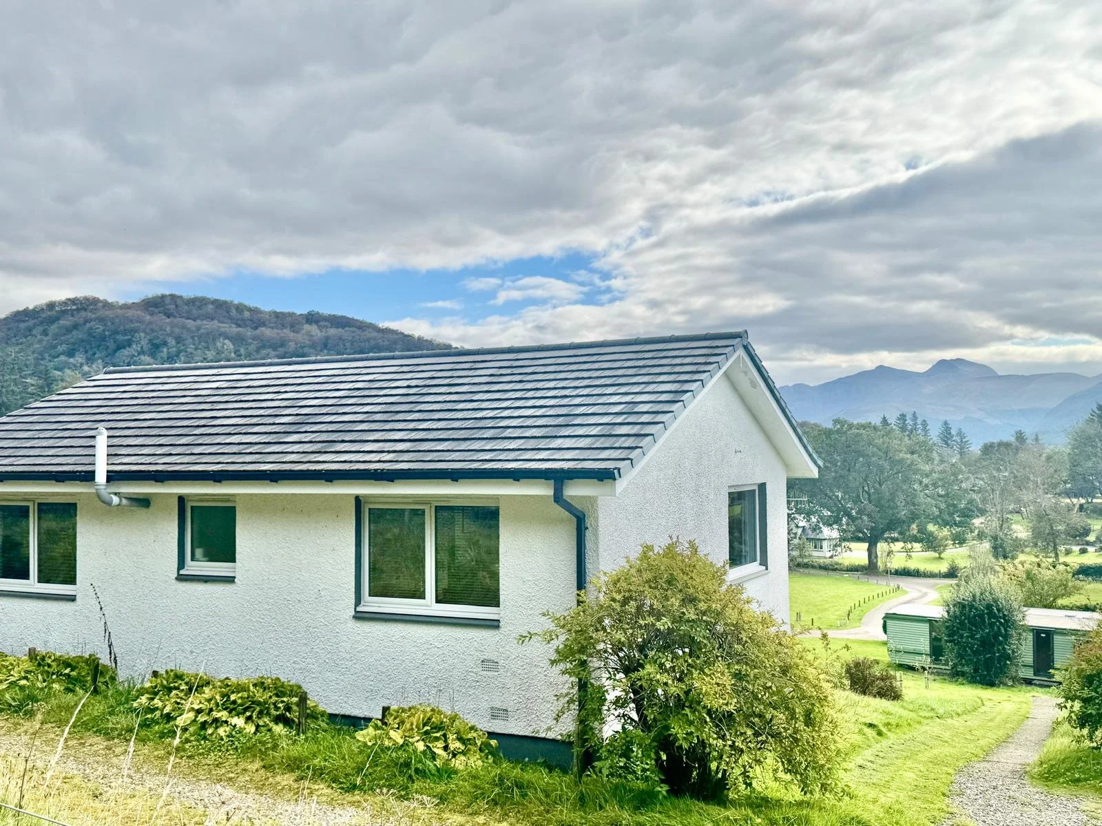 A white house with a gray tiled roof, overlooking a green landscape with trees, bushes, and mountains in the background. The sky is cloudy with patches of blue visible.
