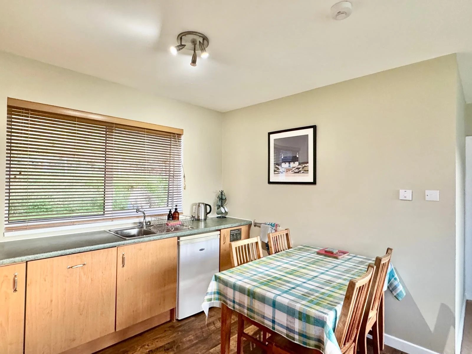 Kitchen with wooden cabinets, a window with blinds, a small fridge, a table with a checked tablecloth, and chairs around it