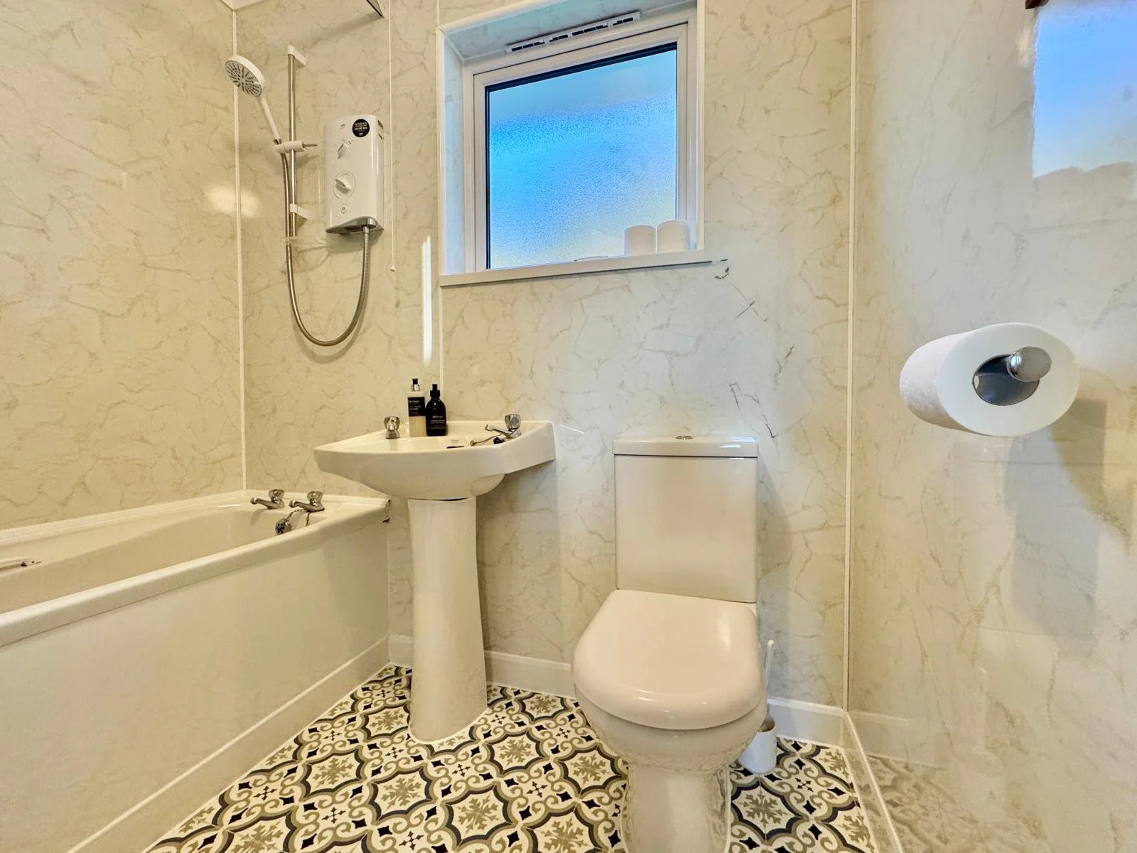 A small vintage-style bathroom with a bathtub, a pedestal sink, a toilet, a window, and patterned tile flooring.