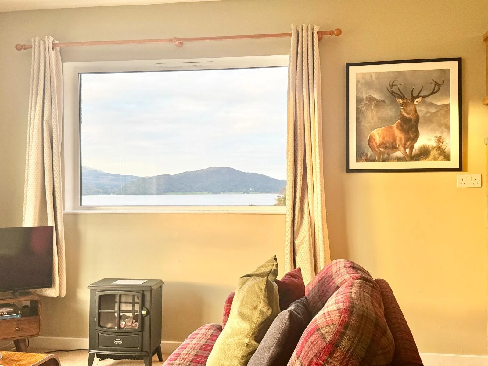 Living room with window showing mountains and water, plaid sofa with pillows, framed deer portrait on wall, small black fireplace, and small wooden TV stand.