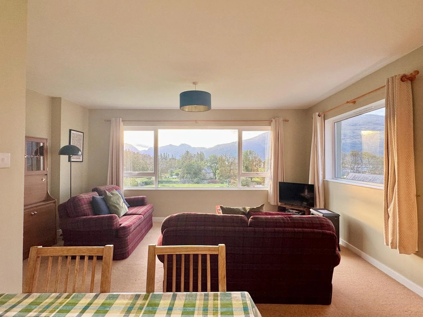 Living room with large window view of mountains, two plaid sofas, a small TV, a wooden cabinet, a floor lamp, and beige curtains.
