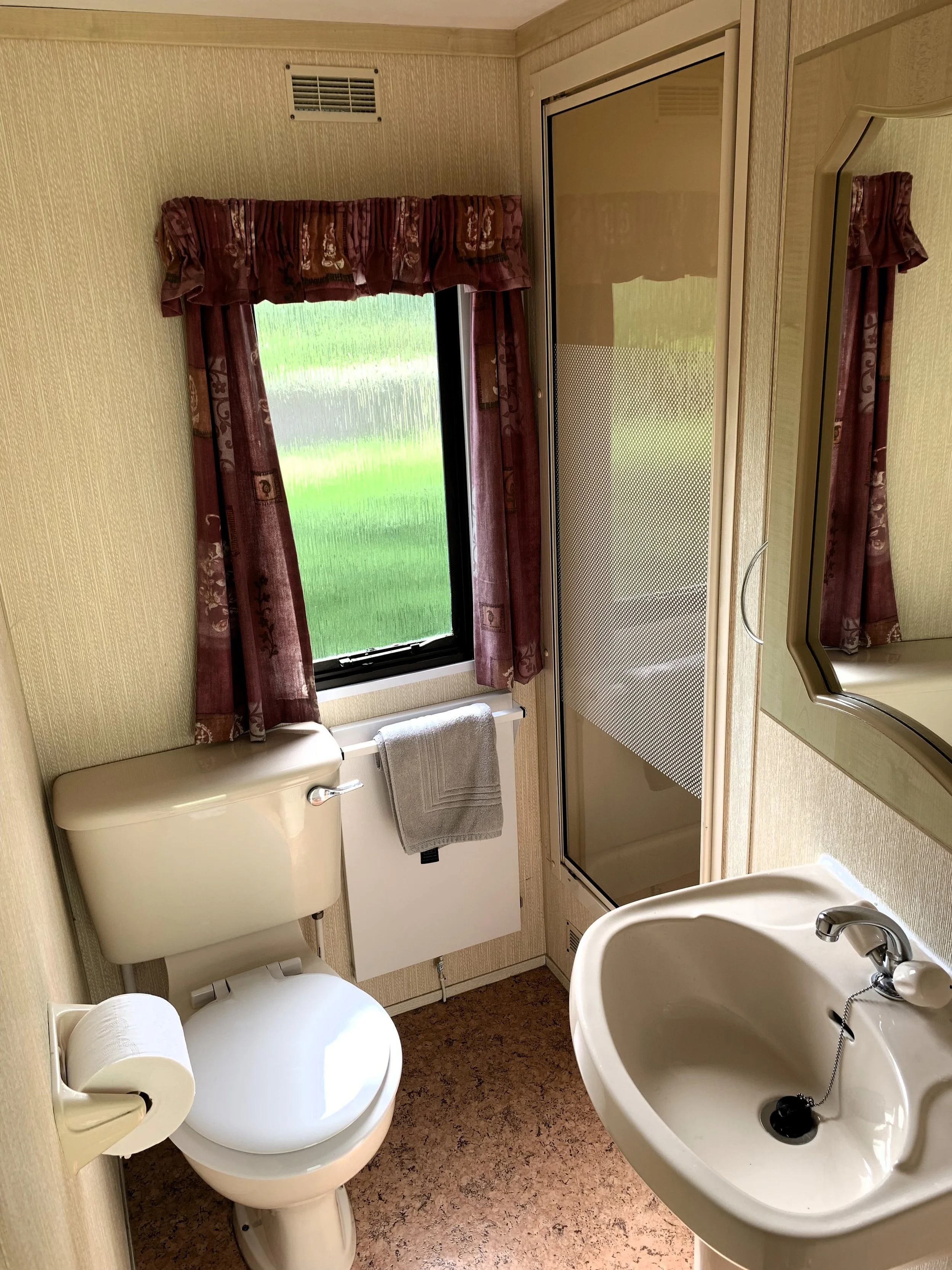 A small bathroom with a toilet, a sink, and a shower stall. There is a small window with red patterned curtains, and a towel hanging on the radiator. The walls are covered with beige textured wallpaper.