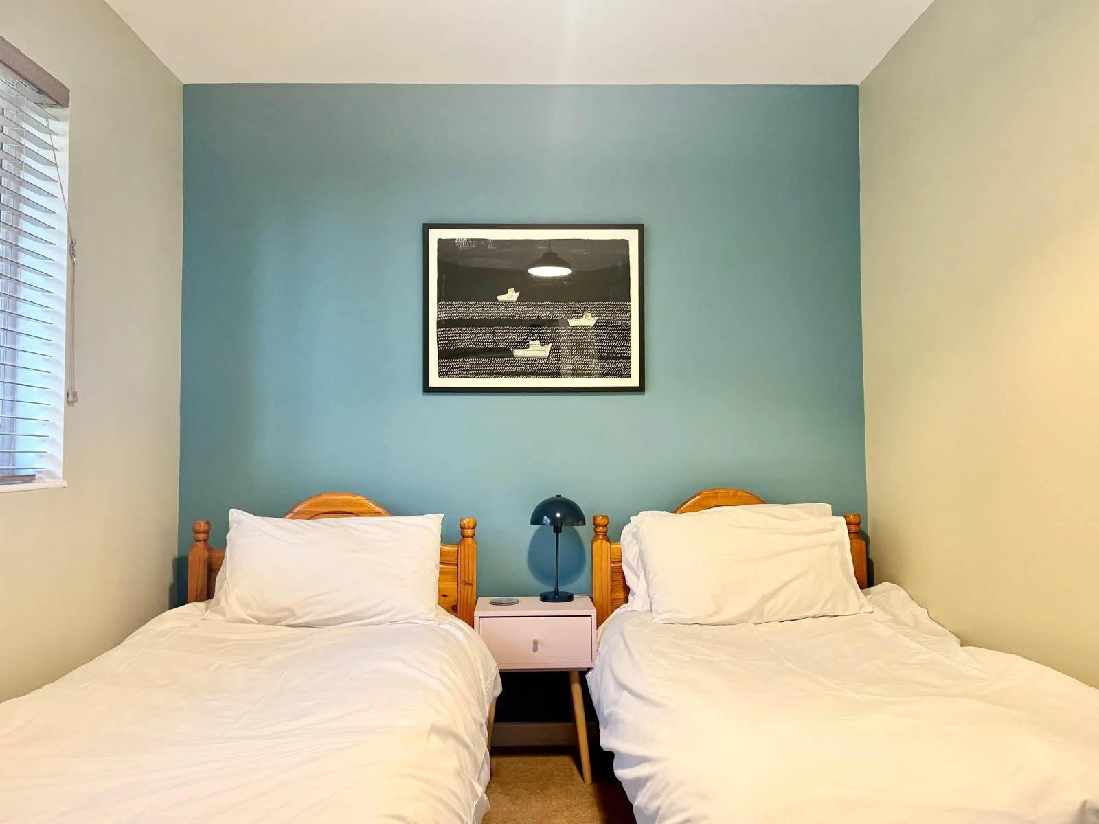 A bedroom with two single beds with white sheets and pillows, a small white nightstand with a blue lamp between the beds, a window with blinds on the left, a framed black-and-white artwork on a blue accent wall above the beds.