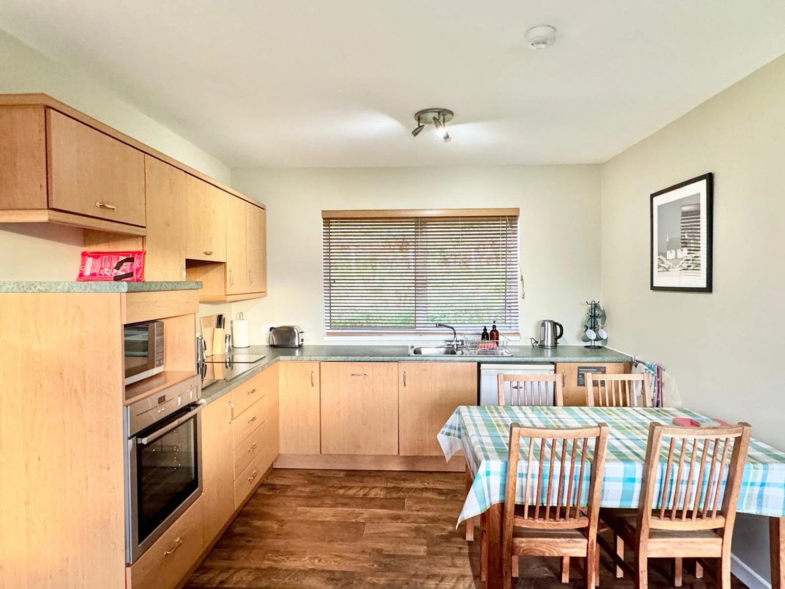 Kitchen with wooden cabinets, a window with blinds, a table with a checkered tablecloth, four wooden chairs, and appliances including a microwave, toaster, kettle, and framed artwork on the wall.