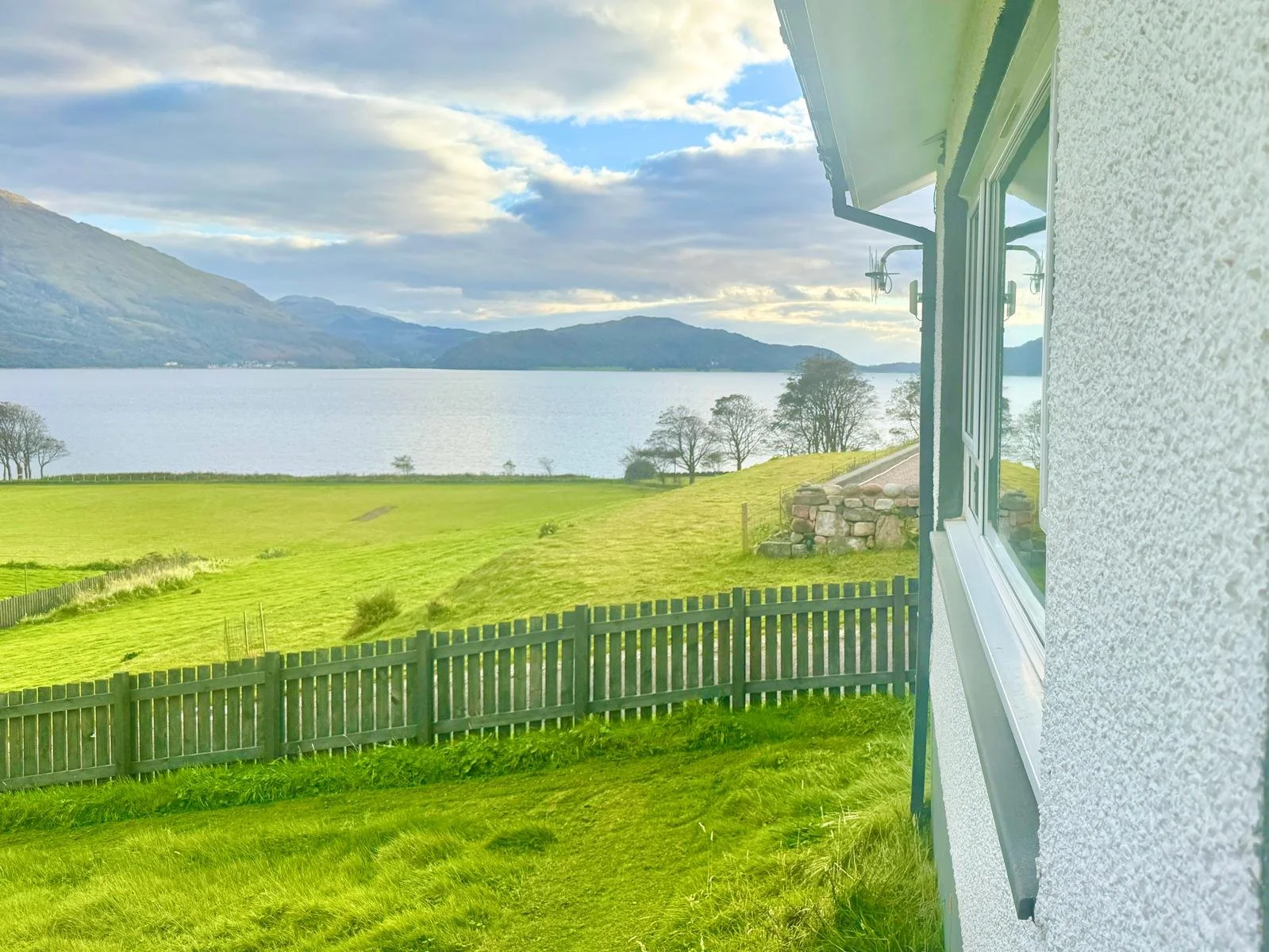 A scenic view of a lake surrounded by green mountains, seen from a house with white walls and a large window, with a green grassy yard and a wooden fence in the foreground.