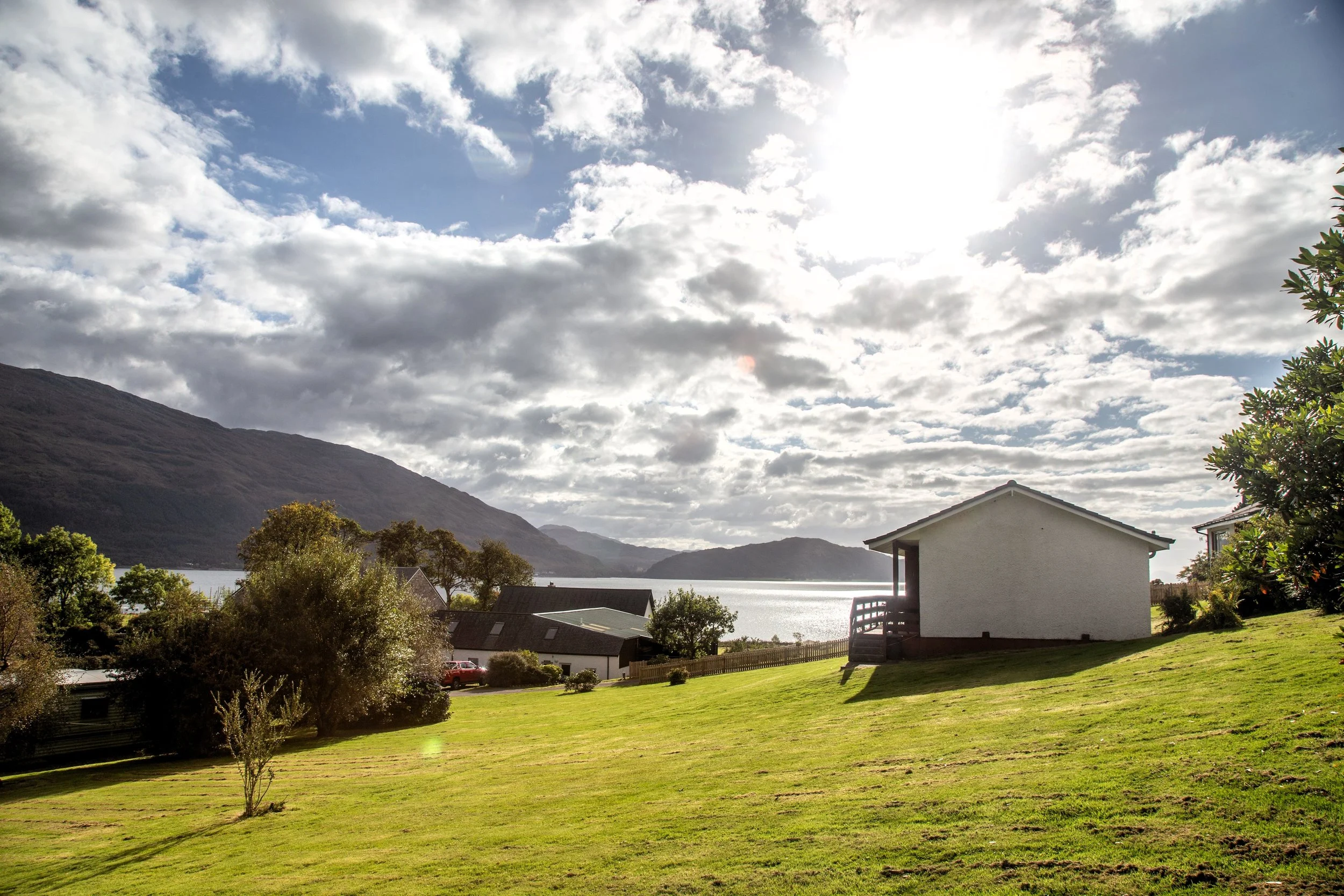 A sunny landscape with a grassy yard, a white house with a porch, and a lake surrounded by mountains in the background, under a partly cloudy sky.