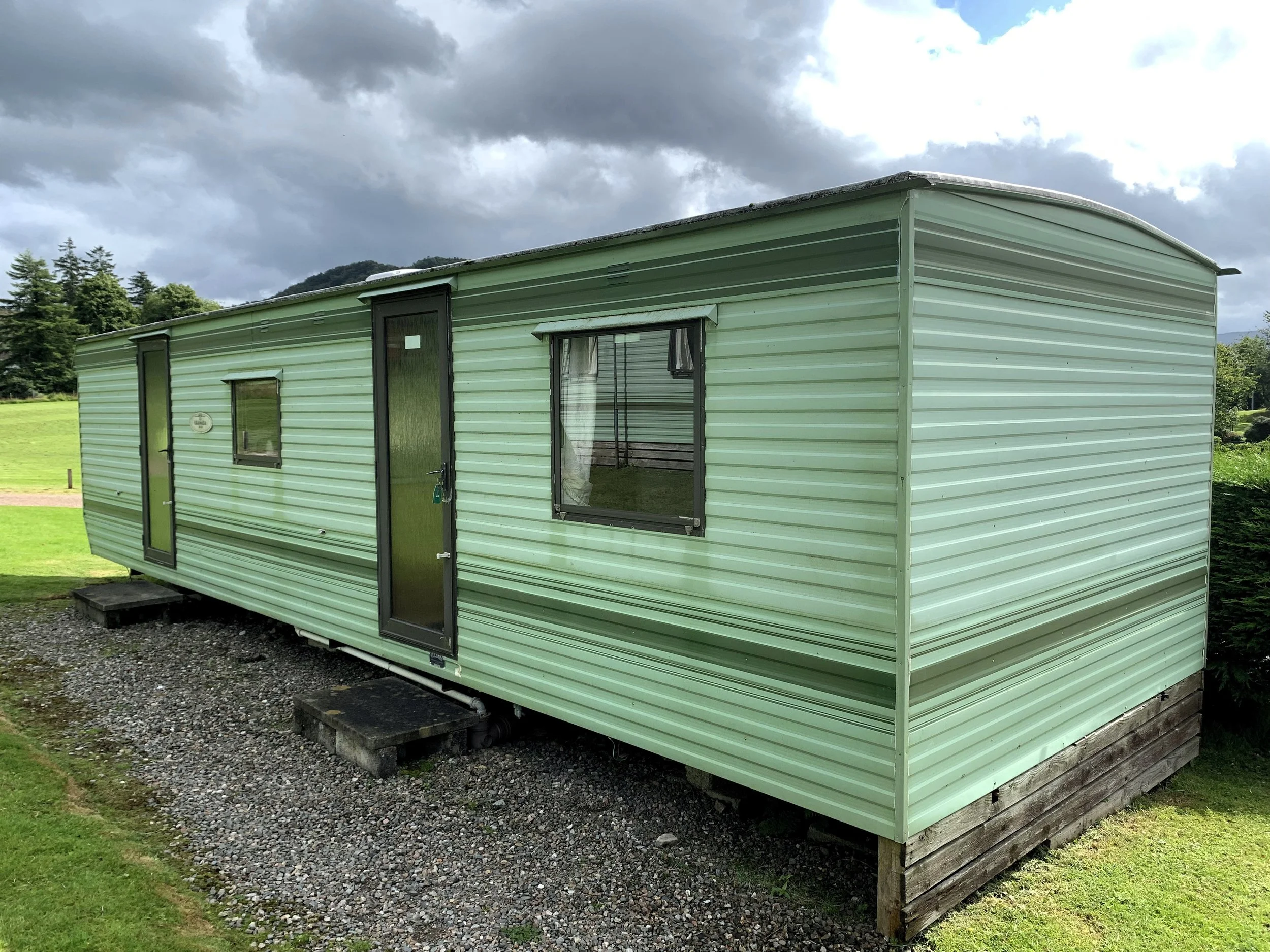 A mint green mobile home with metal siding, situated on a gravel foundation, with three steps leading up to the entrances, surrounded by grassy area and trees, under cloudy skies.