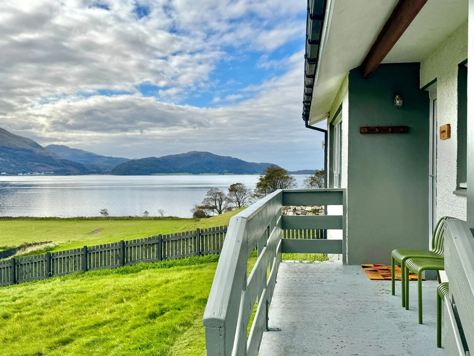 View from a porch overlooking a lake, mountains, and a partly cloudy sky, with green grass and a fence in the foreground.