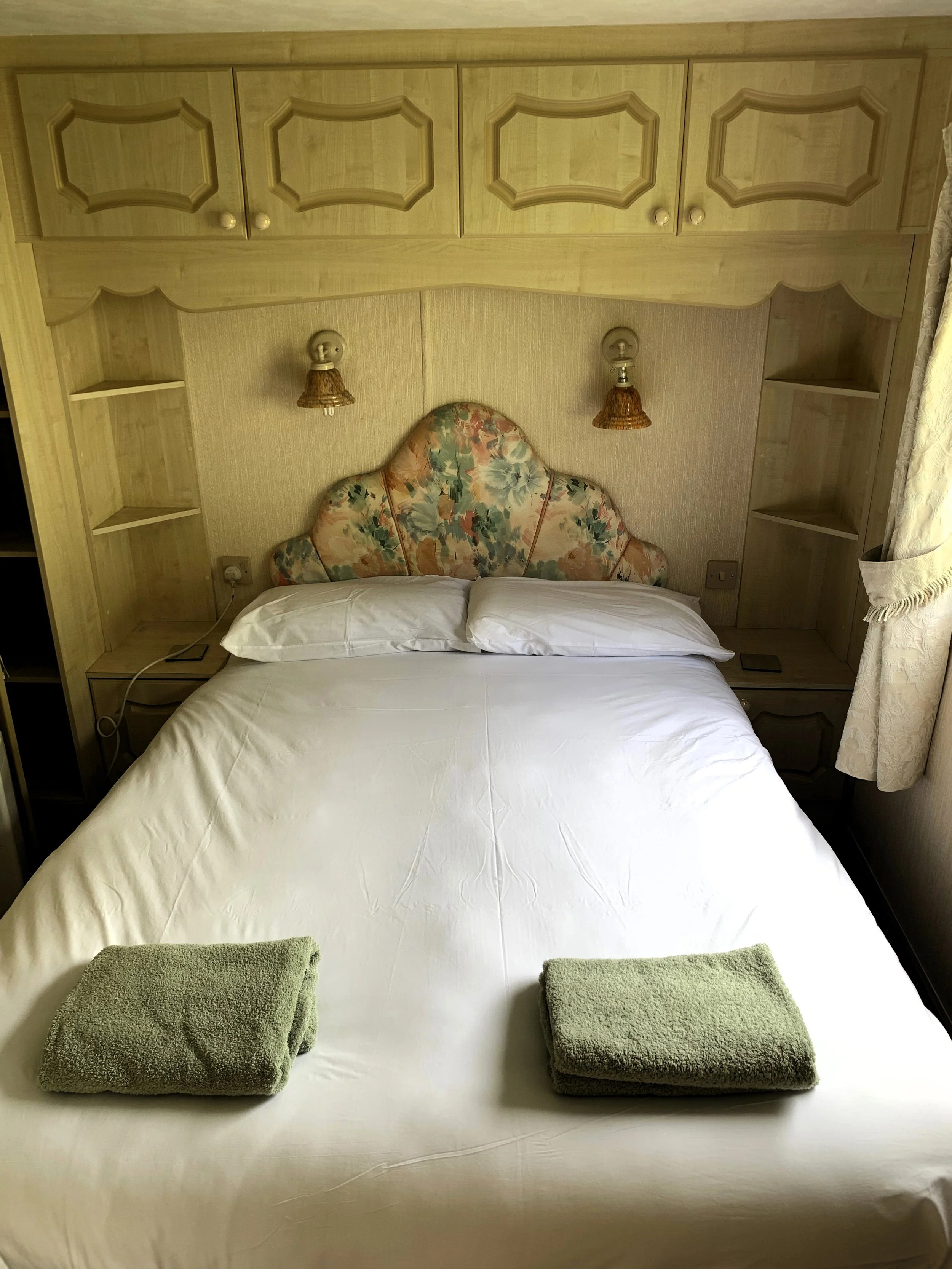 A neatly made bed with two white pillows and two folded green towels on a white sheet, with a floral headboard, flanked by wall-mounted lamps, and wooden cabinetry above the bed, in a cozy bedroom.