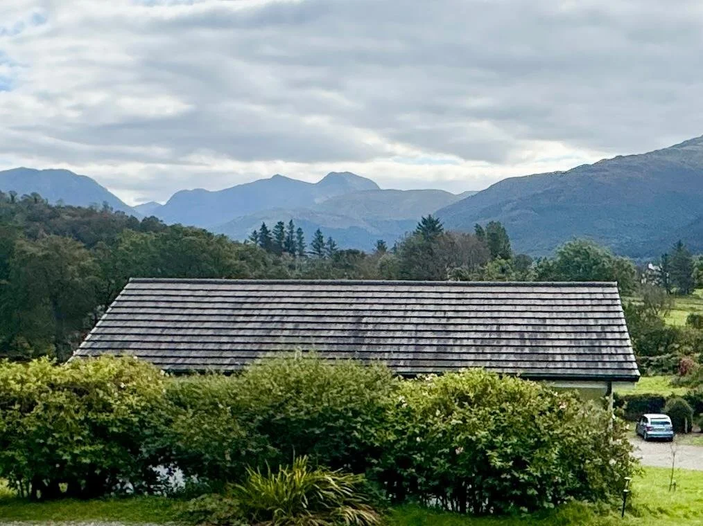 A house with a tiled roof surrounded by bushes, with mountains in the background and a cloudy sky overhead.