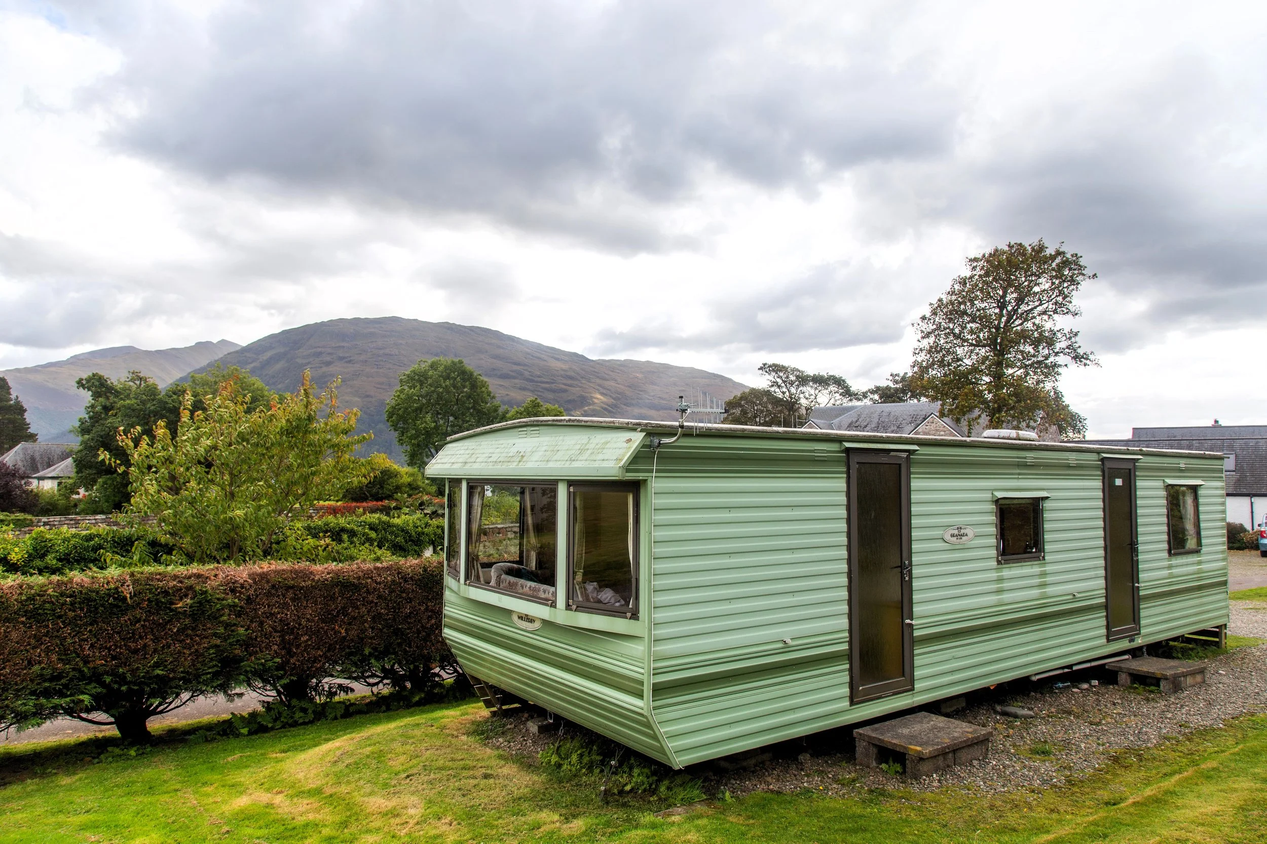 Green mobile home with black doors and windows parked on a grassy area with shrubs, trees, and mountains in the background under cloudy sky.