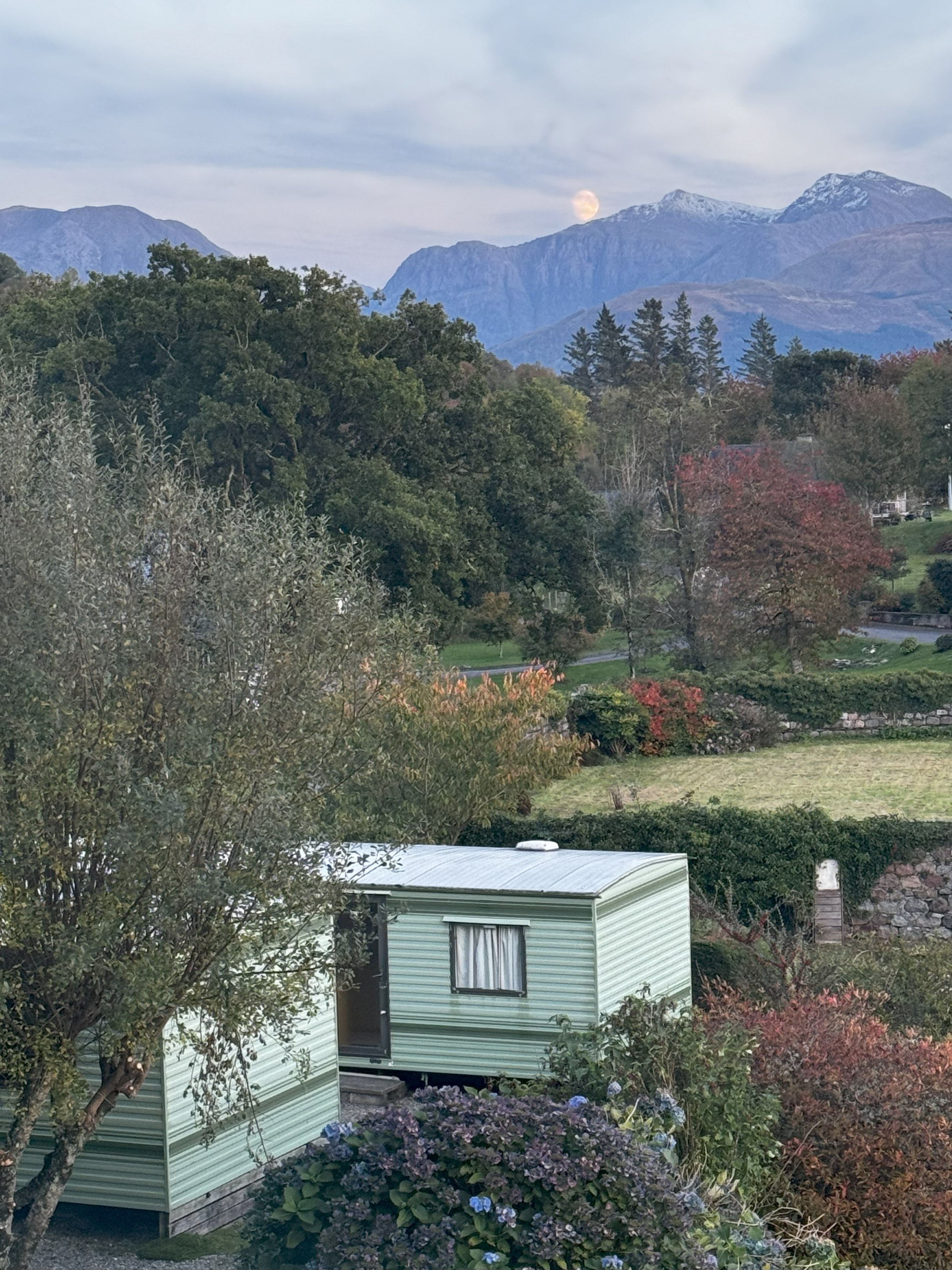 A scenic view of a mountainous landscape with the moon visible near the peaks, a trailer in the foreground, and trees with autumn foliage.