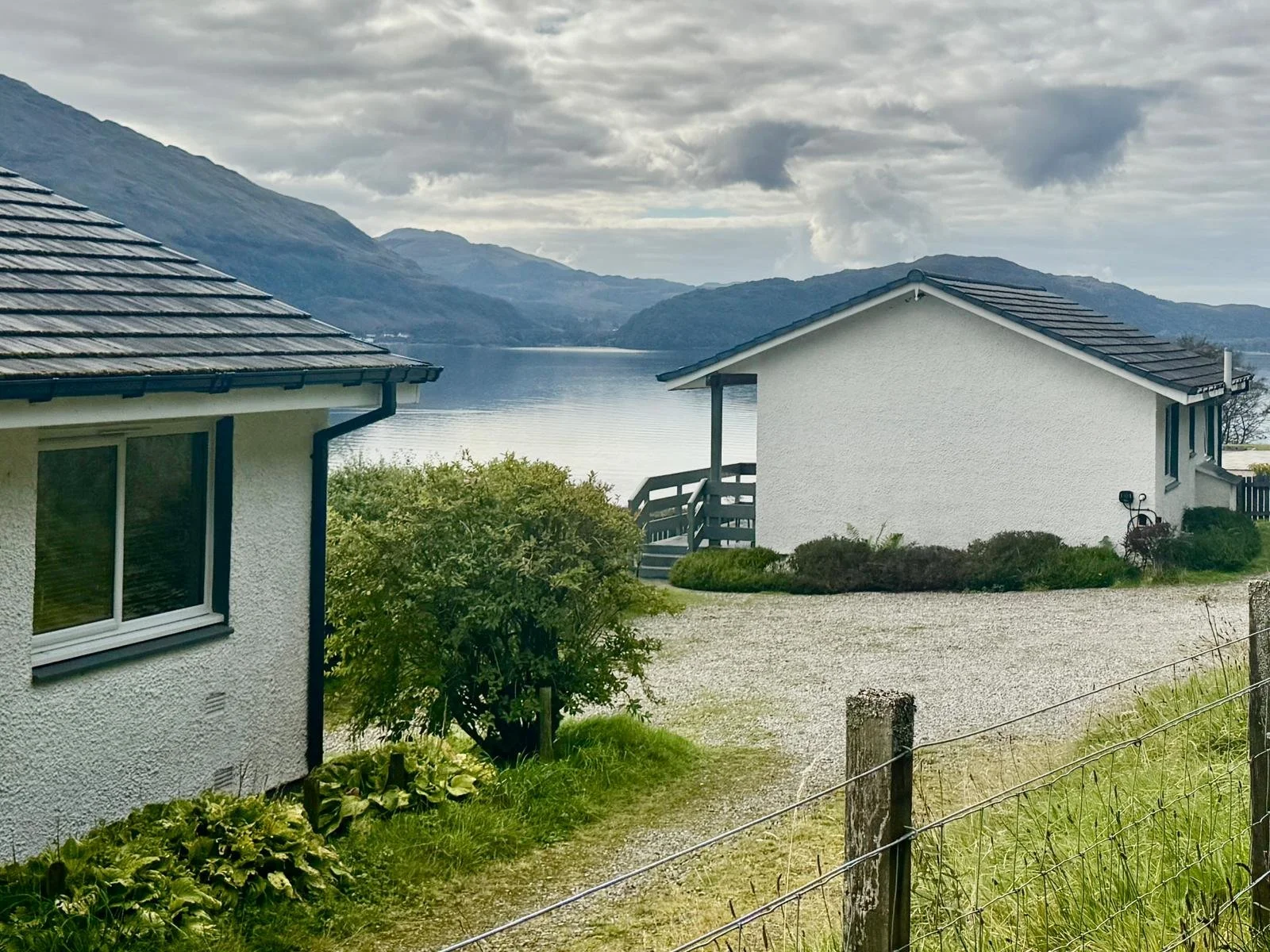 Two white houses on a grassy area by a lake, with mountains in the background and cloudy skies above.