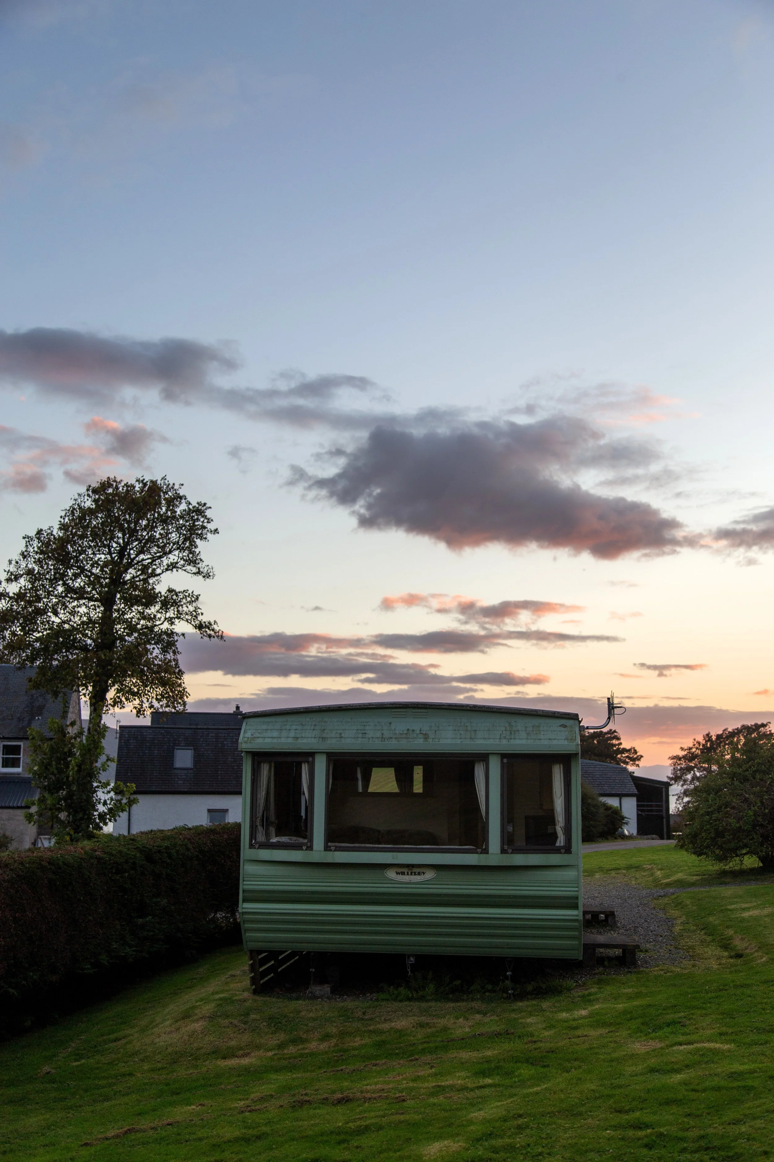 A green caravan parked on a grassy hill at sunset with clouds in the sky and houses in the background.