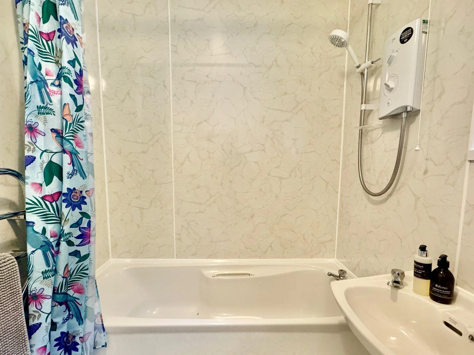 A bathroom with a bathtub, a colorful floral shower curtain, a small sink with bottles, and a showerhead mounted on the wall.
