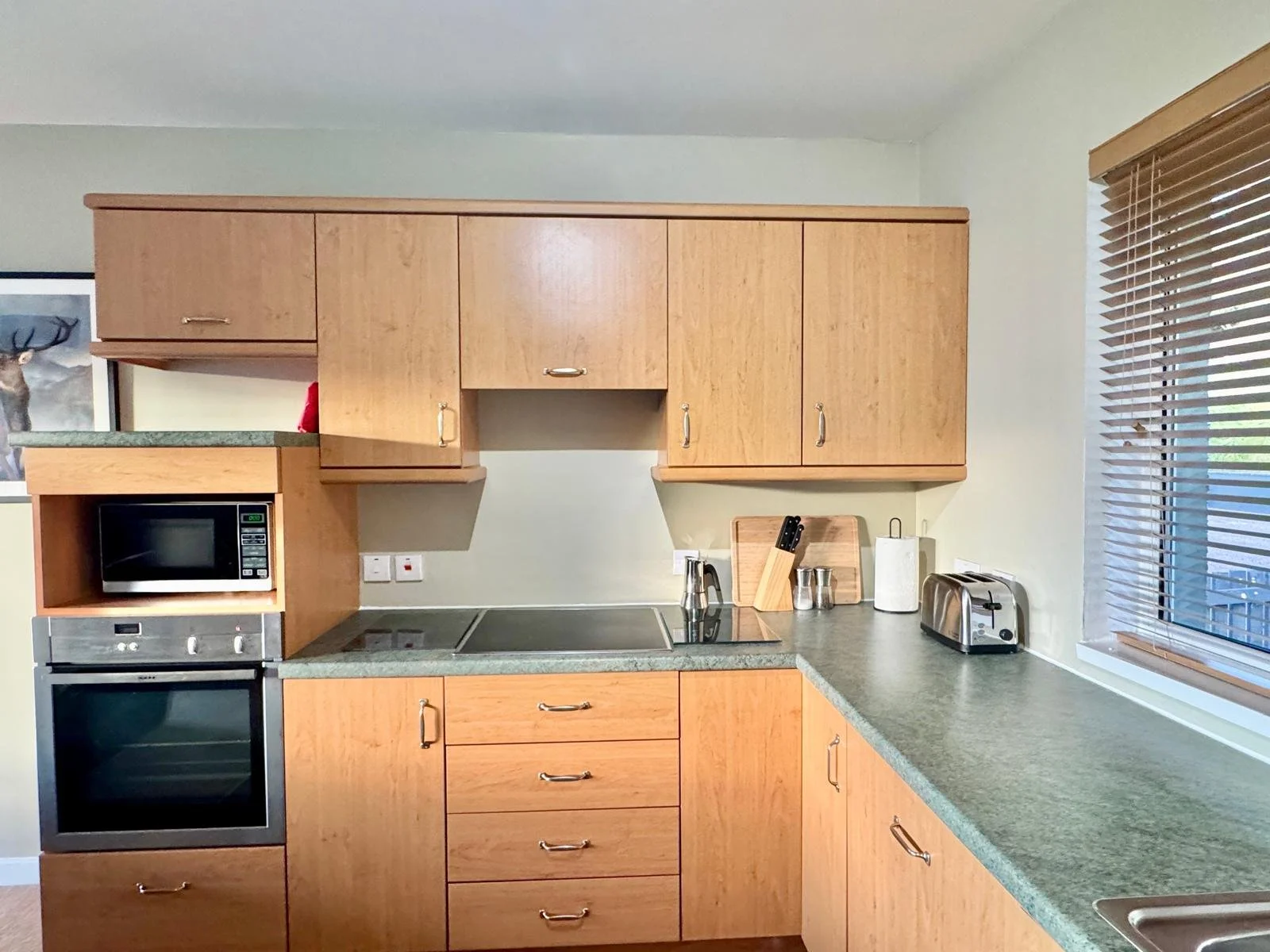 Kitchen with wooden cabinets, a microwave, oven, toaster, and a window with blinds.