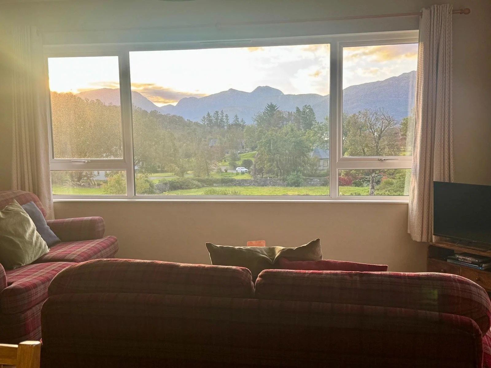 Living room with a large window overlooking a scenic mountain landscape during sunrise or sunset, with a plaid sofa and a small TV on the right.