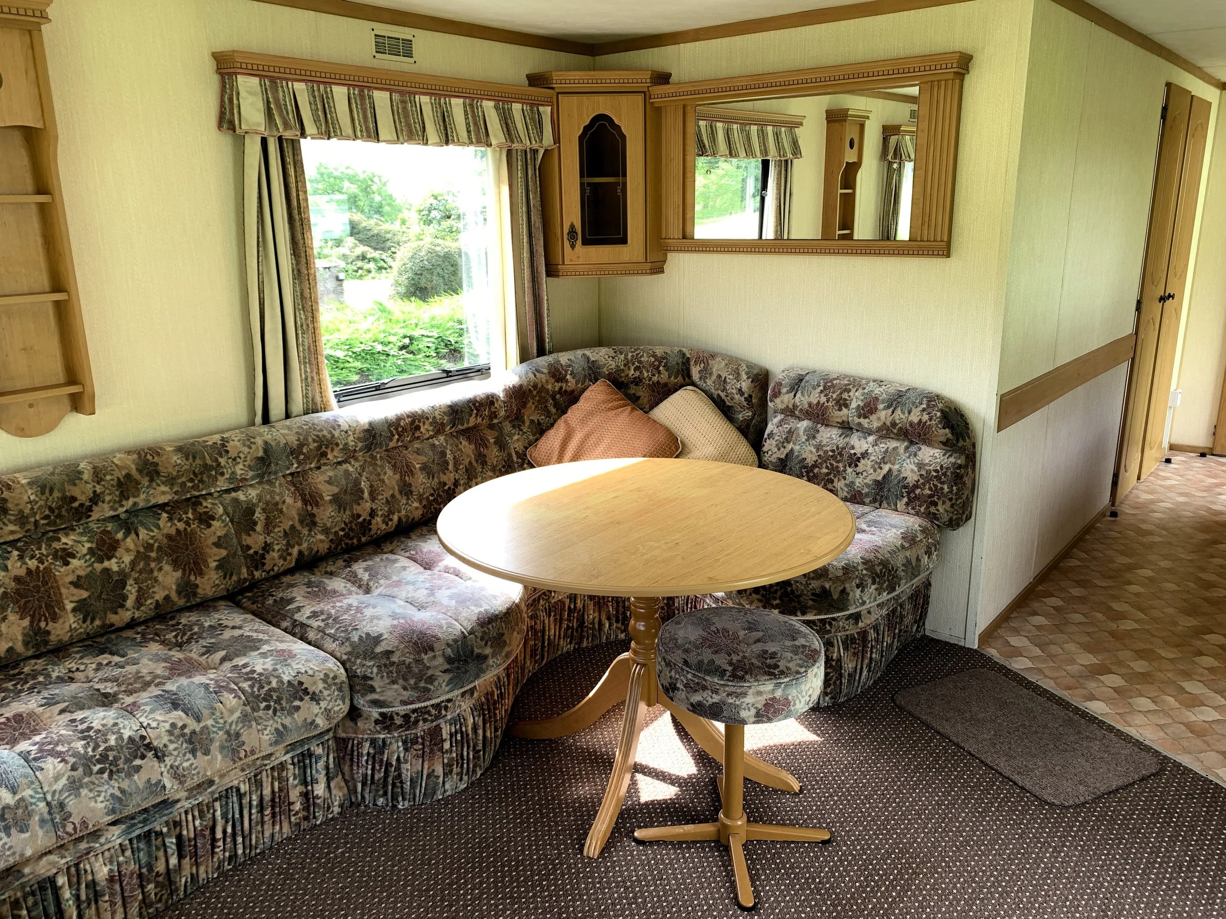 A cozy corner of a living room with a floral upholstered C-shaped sofa, a round wooden table, a small matching stool, and a window with striped curtains showing green foliage outside.
