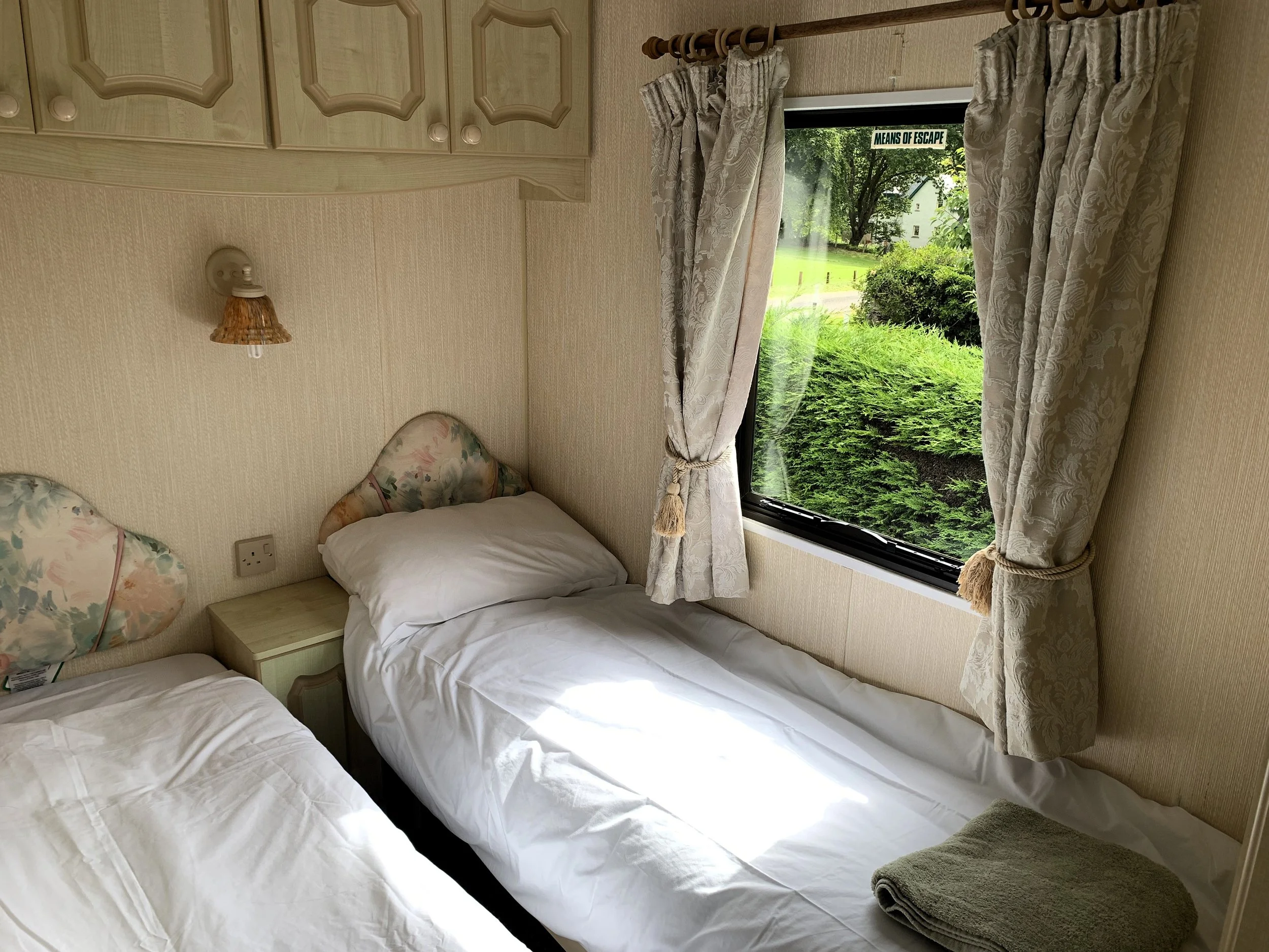 A small bedroom with two twin beds, a window with floral curtains showing a green outdoor scene, and matching upholstered headboards with floral patterns. One bed has a folded towel at the foot, and a small nightstand is between the beds. A wall-moun