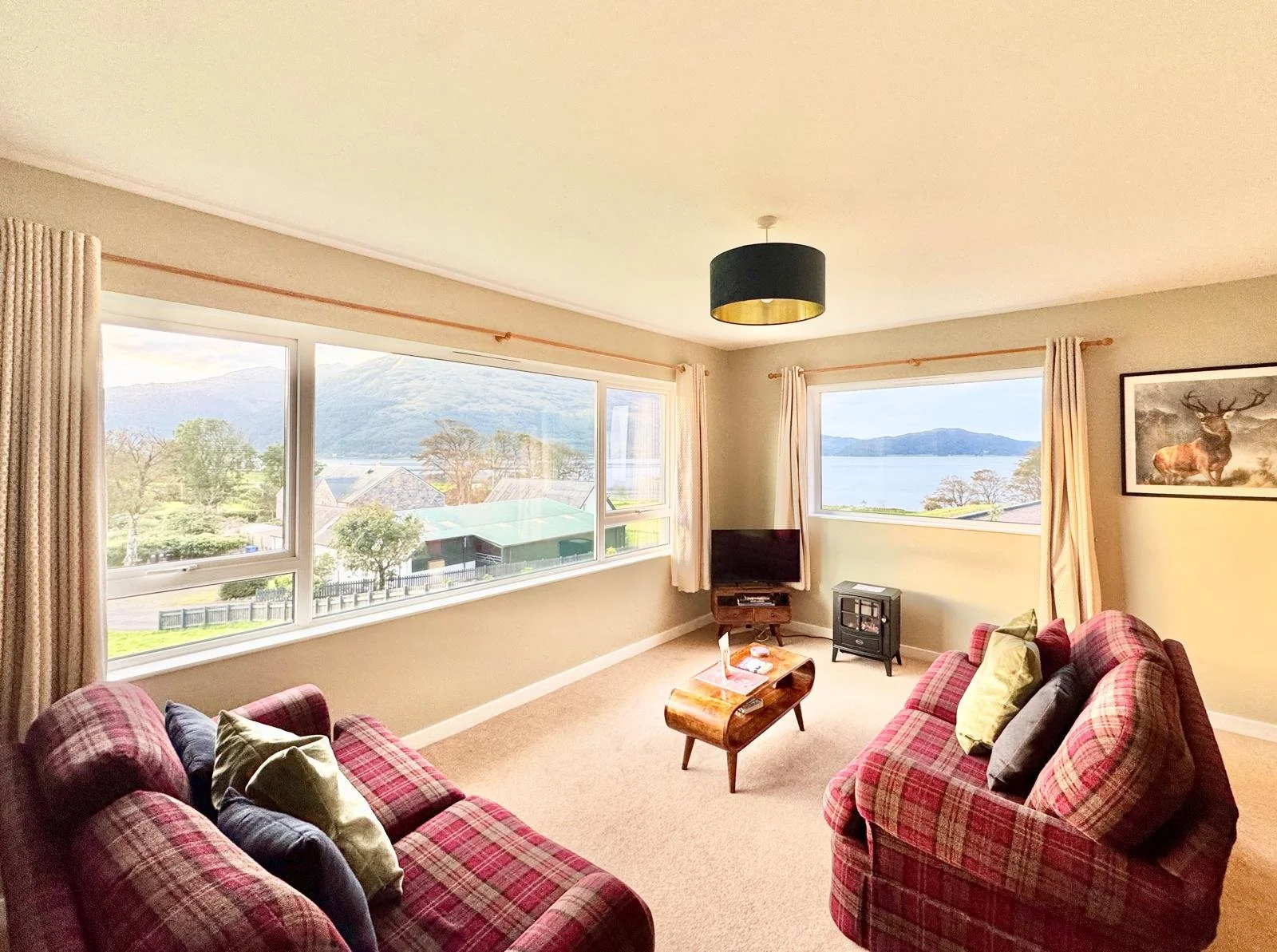 Living room with two pink plaid sofas, cushions, a wooden coffee table with magazines, a small TV, a landscape painting of a deer, large windows showing mountains, trees, water, and a cloudy sky, beige curtains, and a black ceiling light fixture.