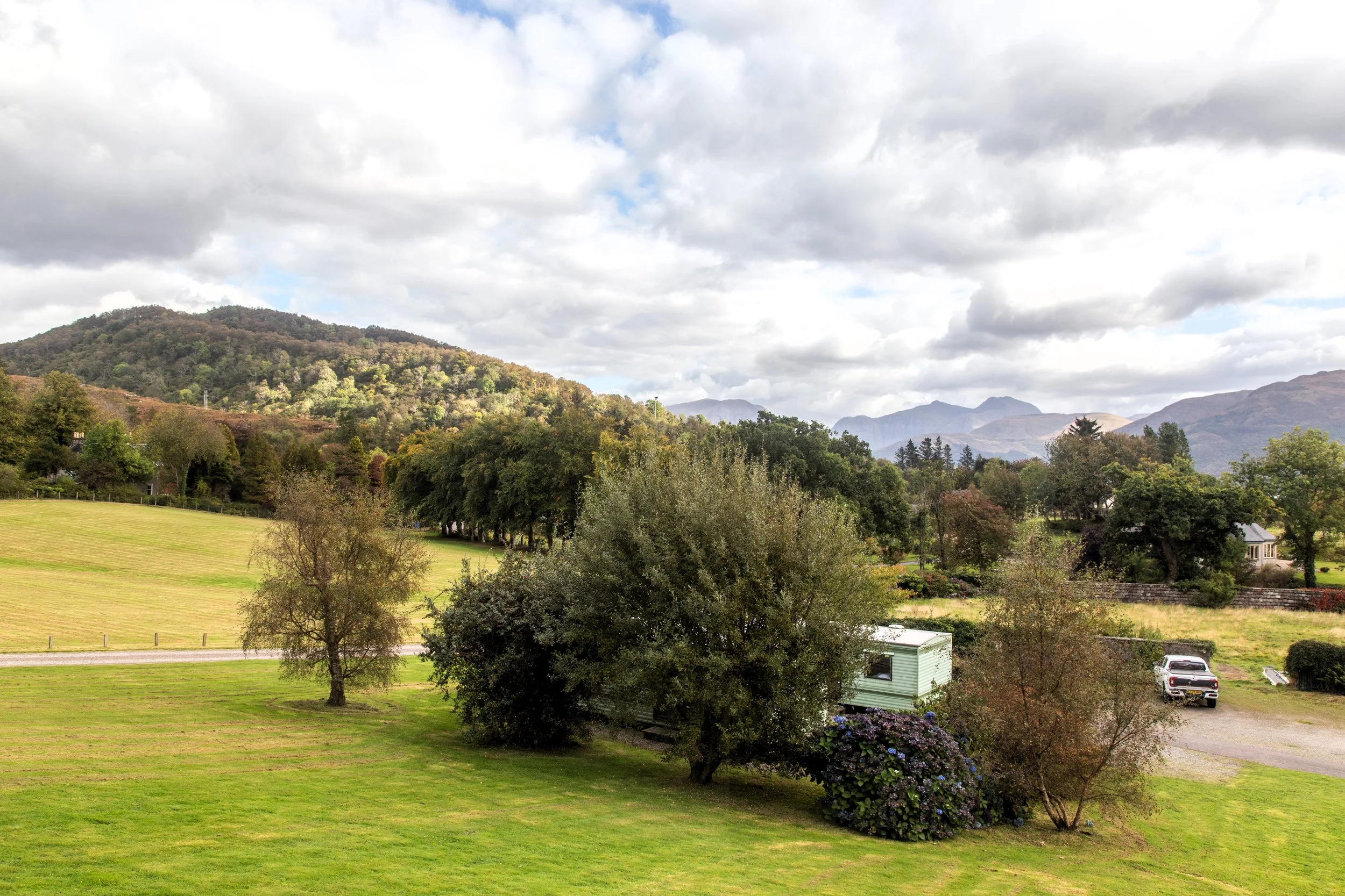 Scenic landscape with rolling green lawns, trees, a white trailer, parked vehicles, and mountains in the background under partly cloudy skies.