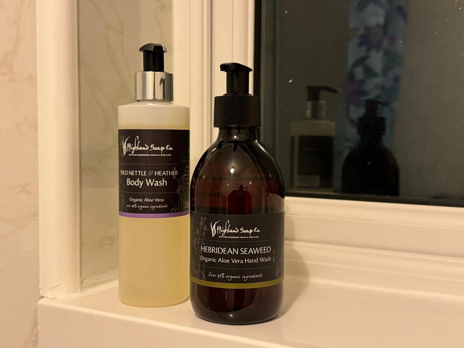 Two bottles of organic aloe vera soap and hand wash on a bathroom windowsill, one labeled 'Wild Nettles & Heather Body Wash' and the other labeled 'Hebridean Seaweed Hand Wash.'