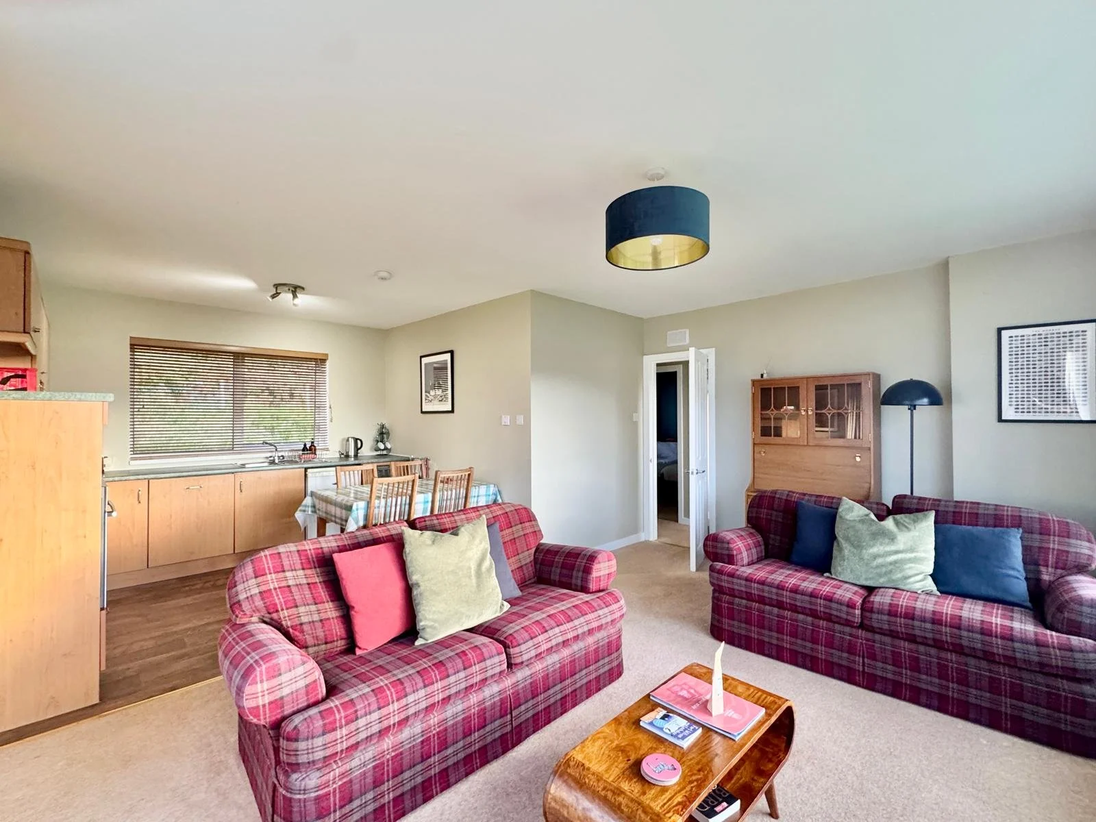 Living room with two plaid sofas, throw pillows, a wooden coffee table with magazines, a background kitchen with a dining table, window blinds, and a doorway to another room.