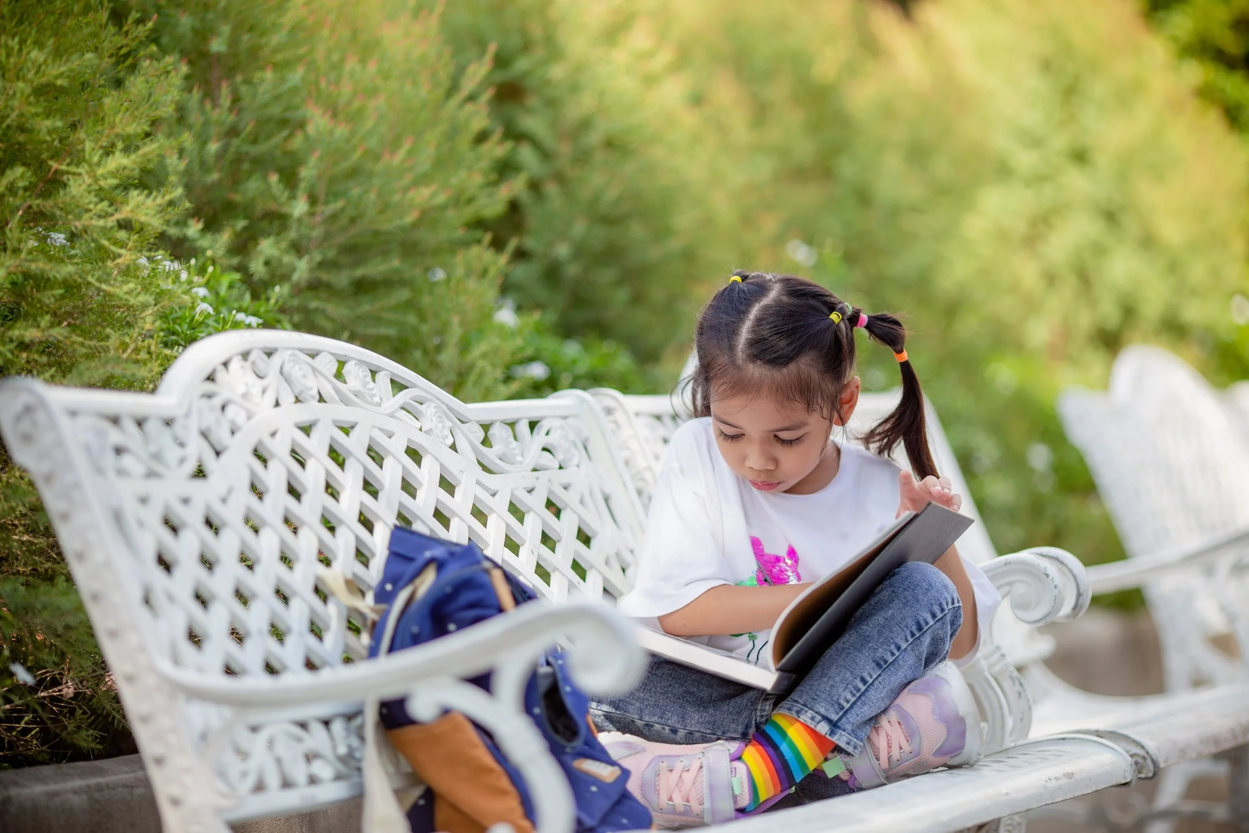 Small child reading a book on a bench in a park. She has pigtails and is wearing rainbow-striped socks.
