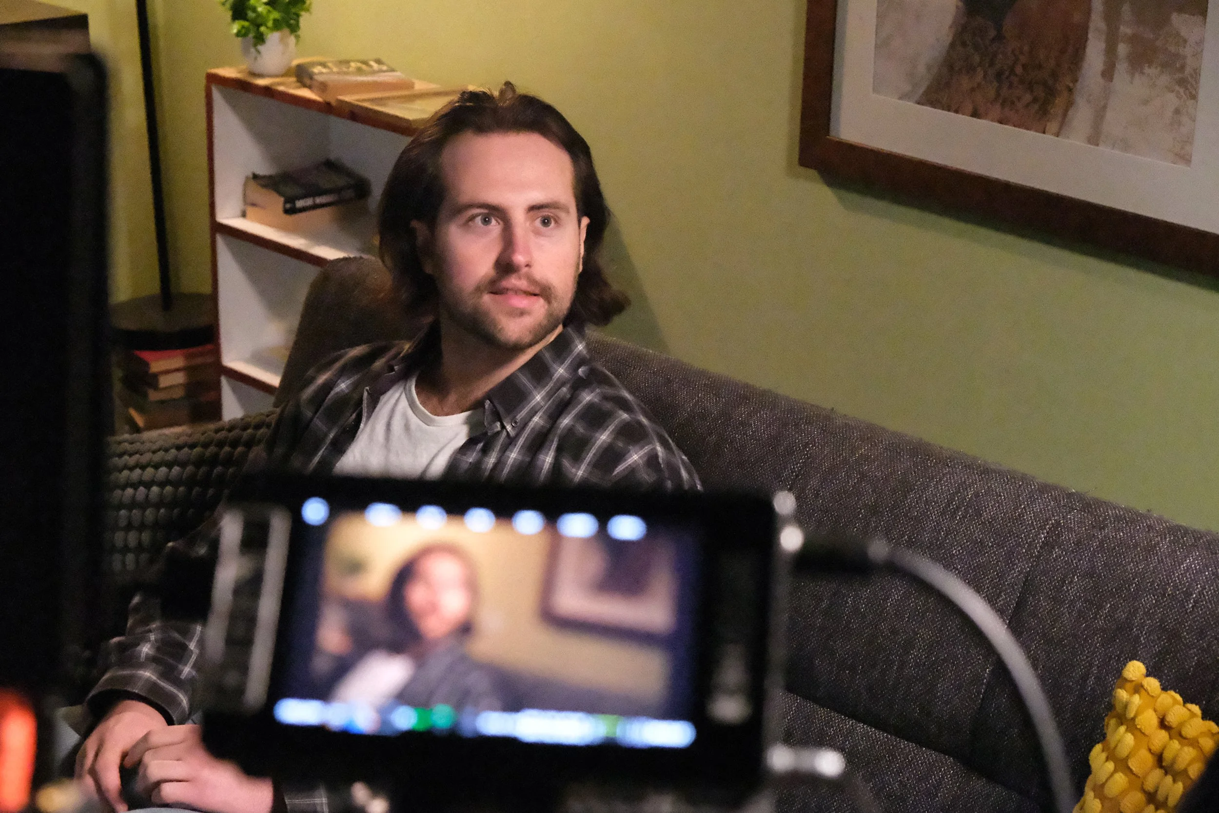 A man with dark hair, beard, and plaid shirt sitting on a couch, looking slightly to his right, with a camera or phone capturing his image in the foreground.
