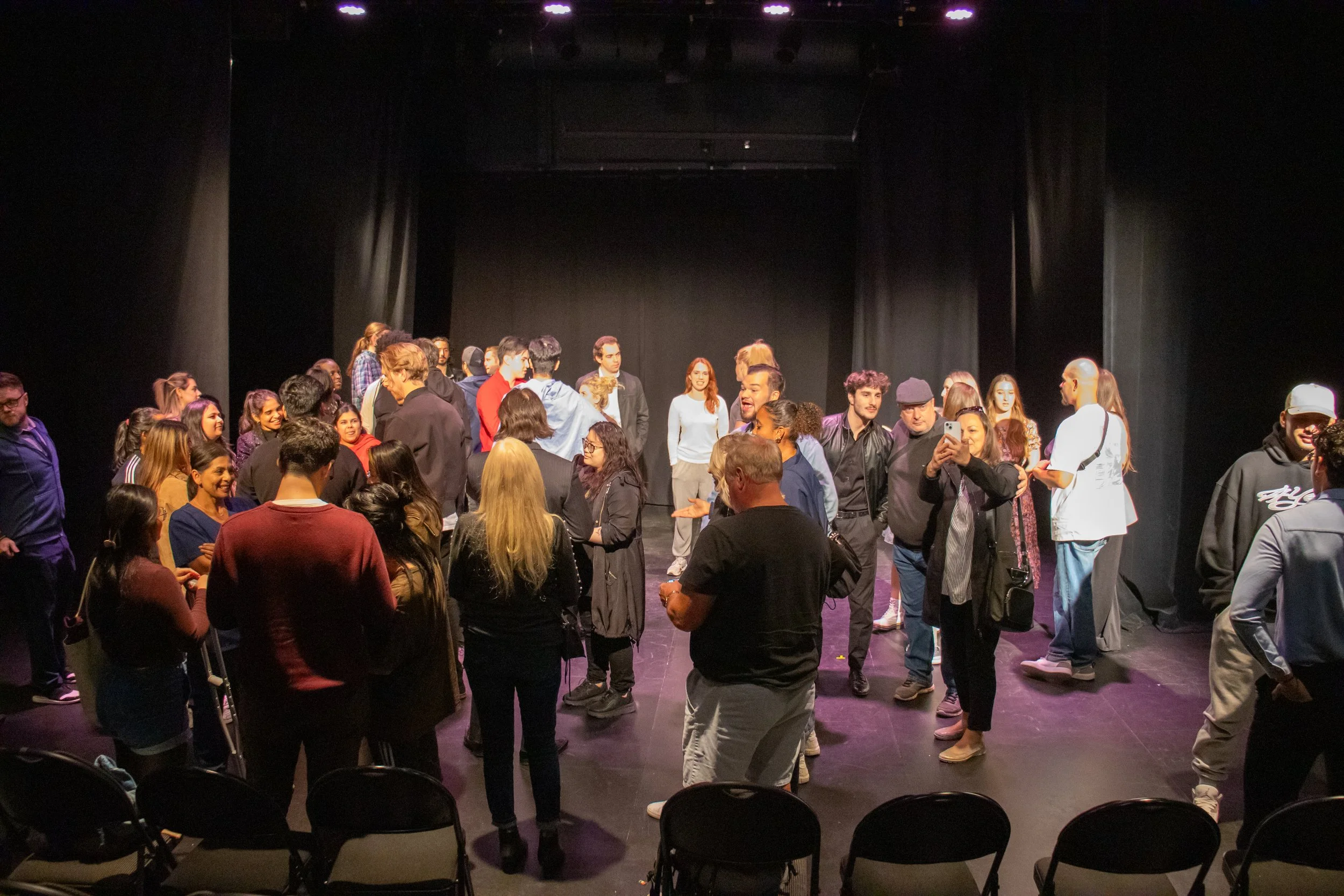 People gathered on a stage, some talking, and one person taking a photo with a smartphone, with black curtains and stage lights overhead.