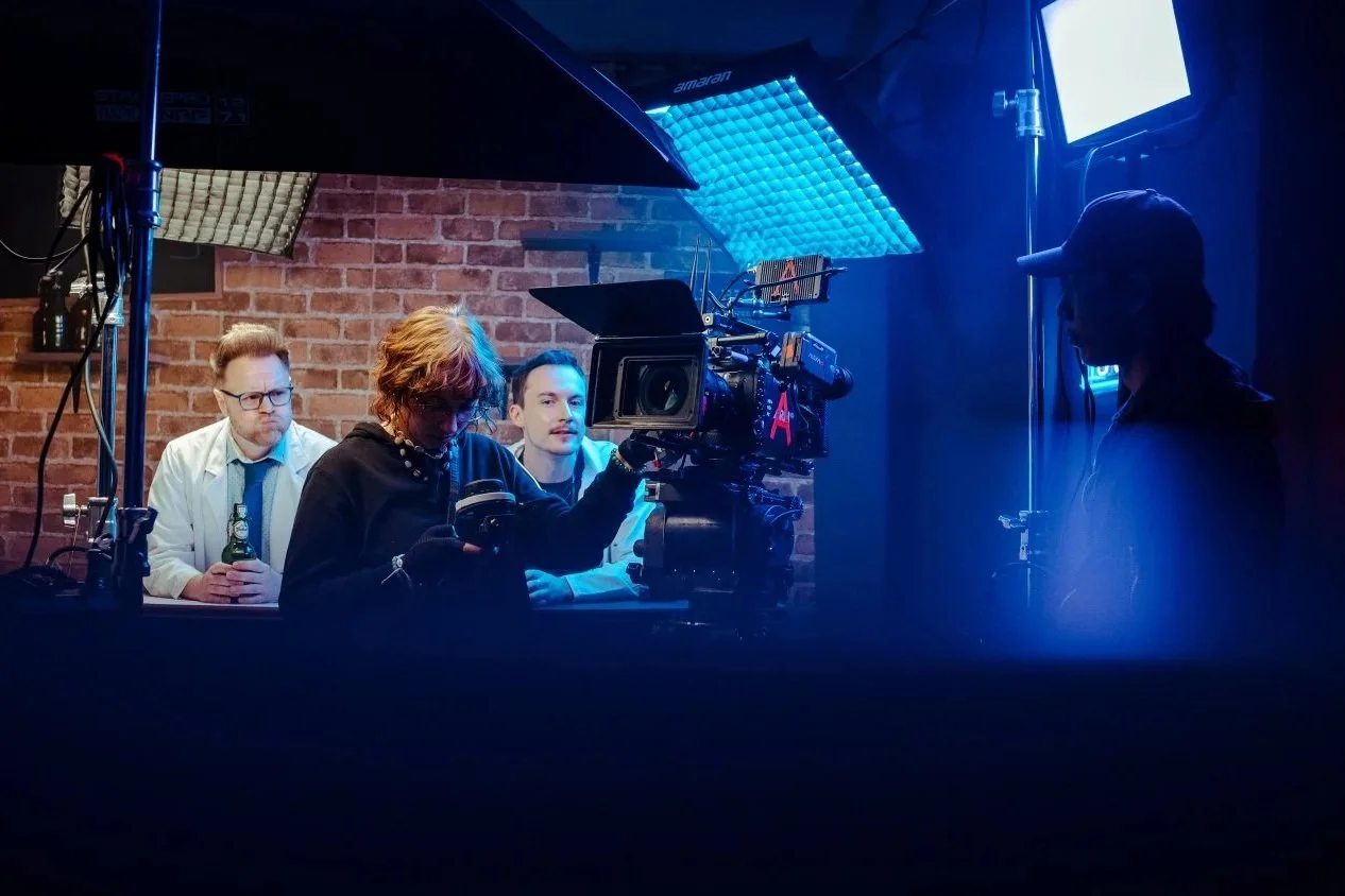People filming a scene in a studio with professional camera equipment and lighting, against a brick wall background.