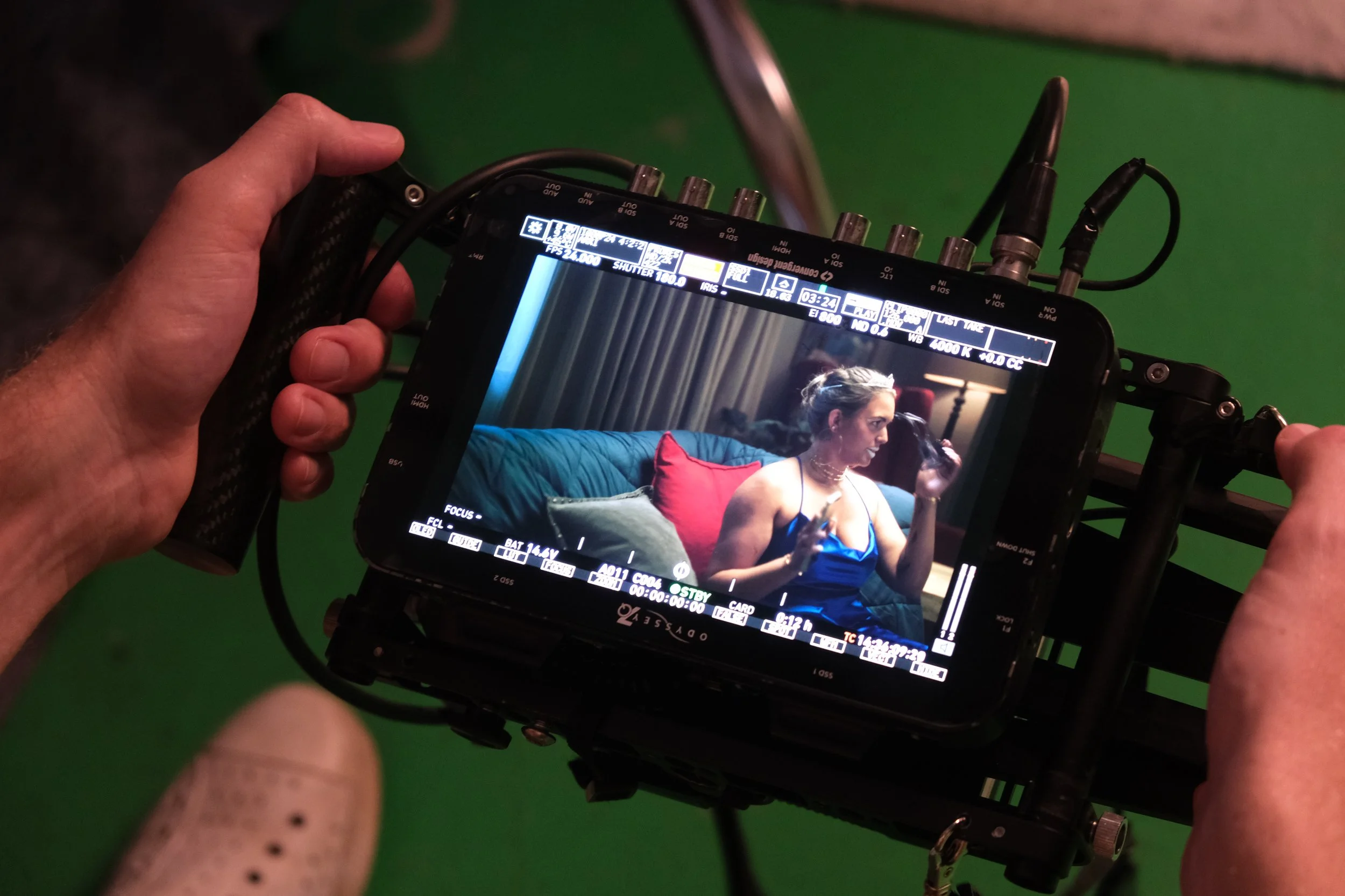 Close-up of a monitor showing a woman in a blue dress sitting on a green couch with red and green pillows, holding a glass, with studio setting in the background.