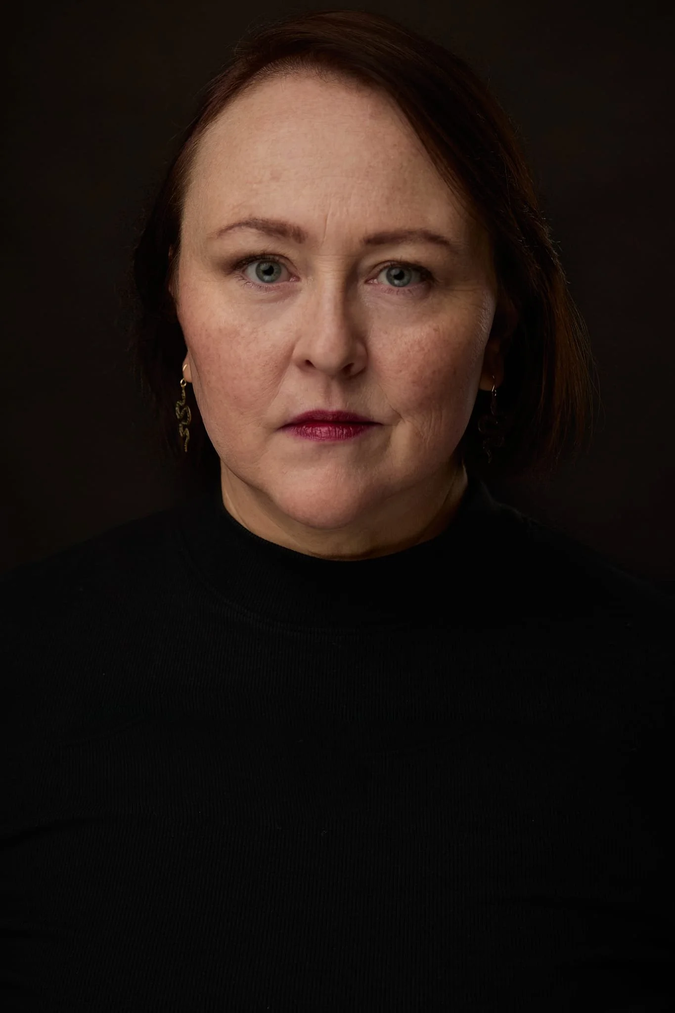 Portrait of a woman with short auburn hair, blue eyes, wearing a black turtleneck and gold earrings, against a dark background.