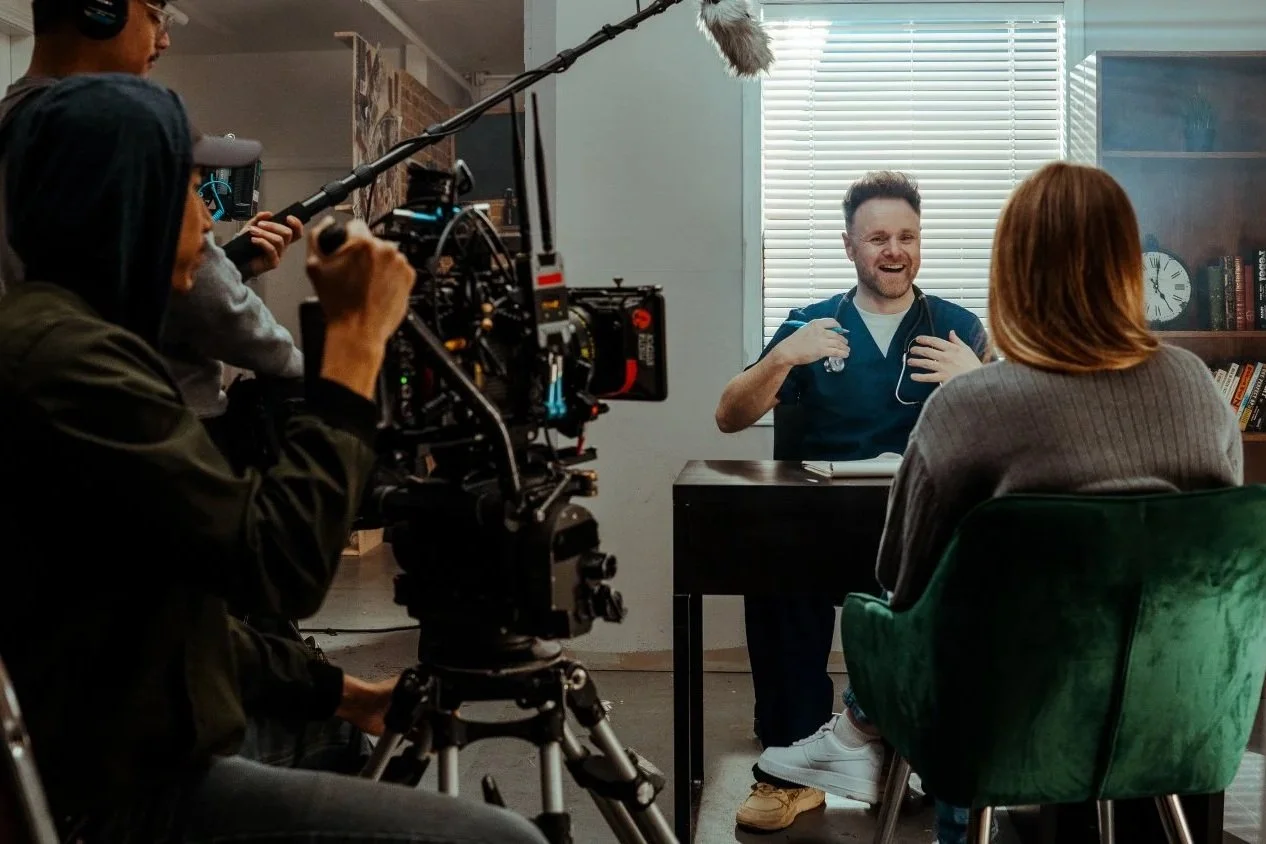 A man in medical scrubs being filmed during an interview or conversation in an indoor setting with a window and bookshelf.