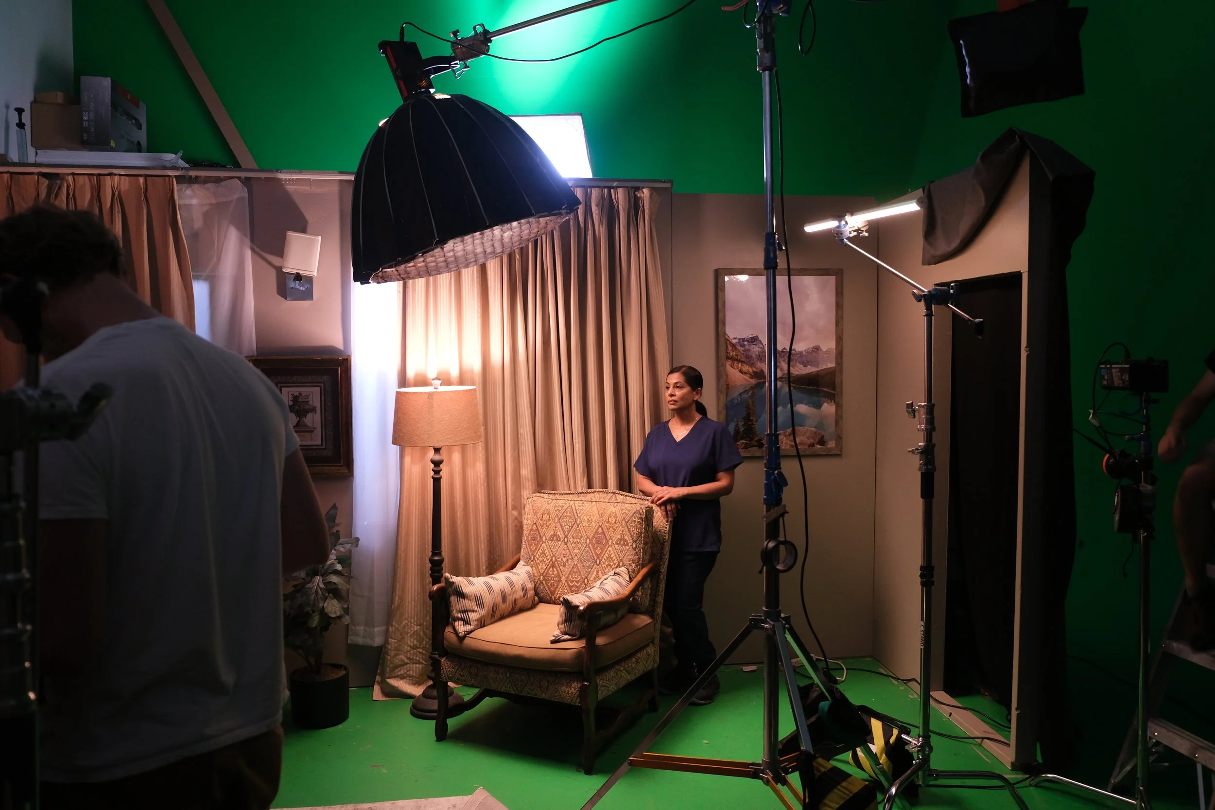 A woman standing in front of a camera on a filming set with a backdrop of a sofa, lamp, curtains, and landscape artwork, surrounded by studio equipment including lights and green screens.