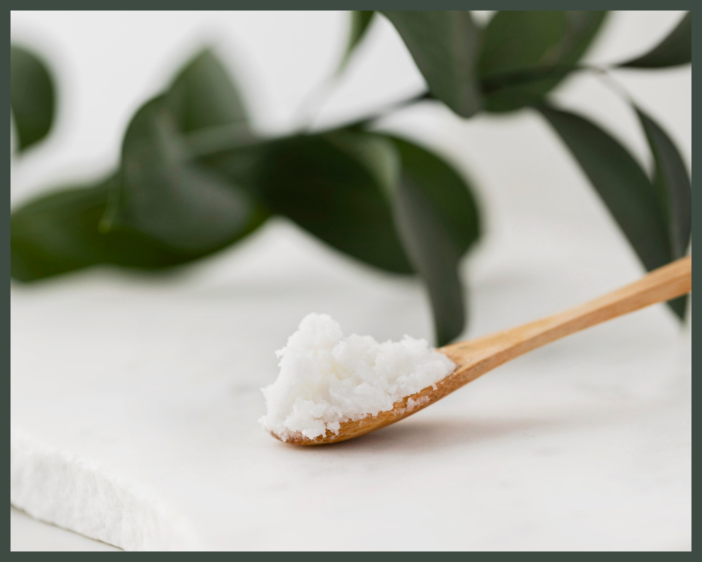 A wooden spoon holding a small amount of white coconut oil on a marble surface with green leaves in the background.