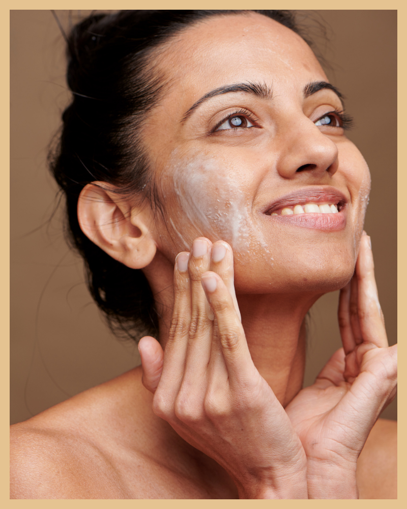 Person applying skincare product on face, smiling with eyes looking upwards, brown background.