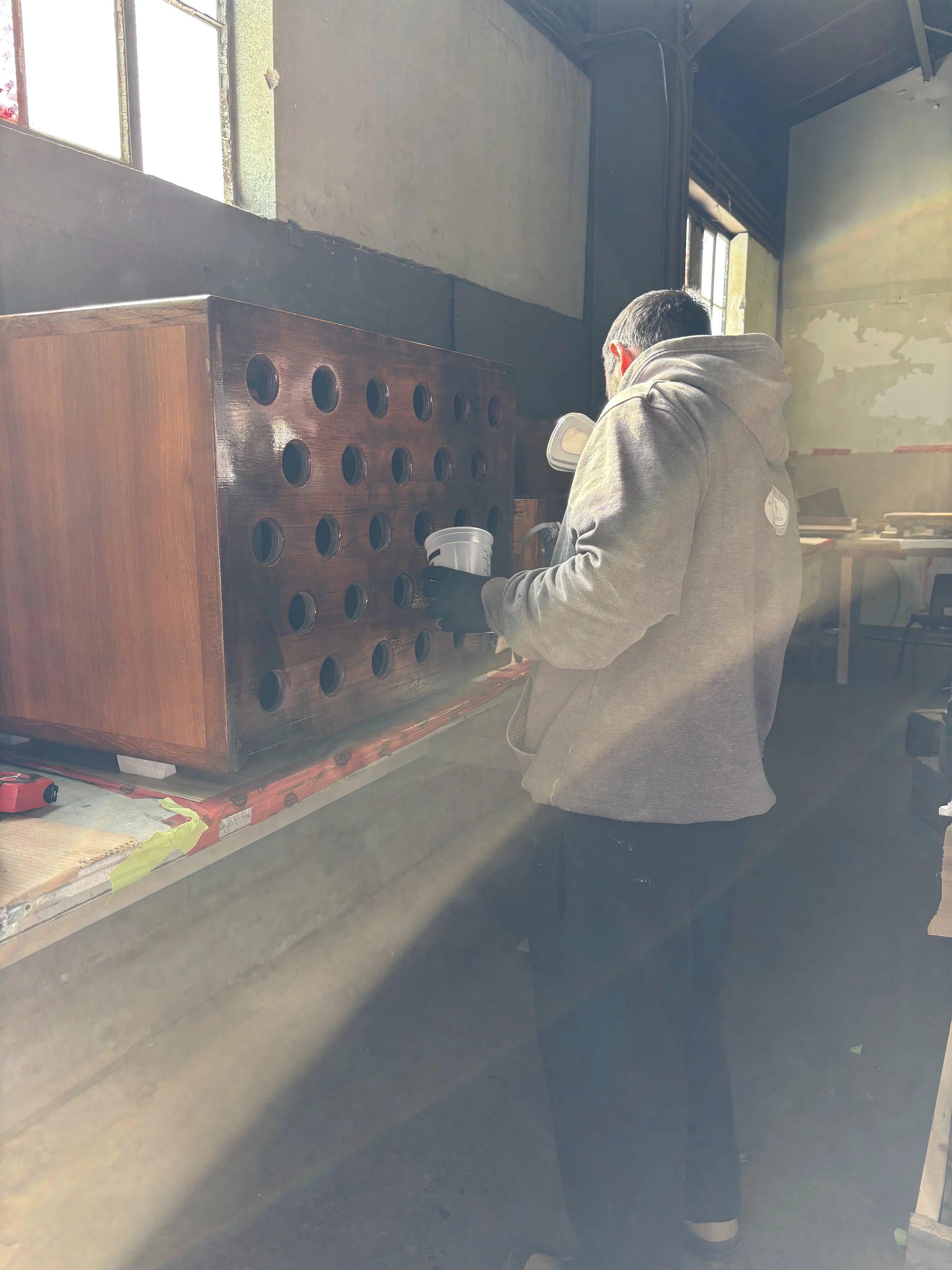 A person wearing a gray hoodie and black gloves is working on a wooden piece with multiple circular holes in a workshop with large windows and sunlight streaming in.