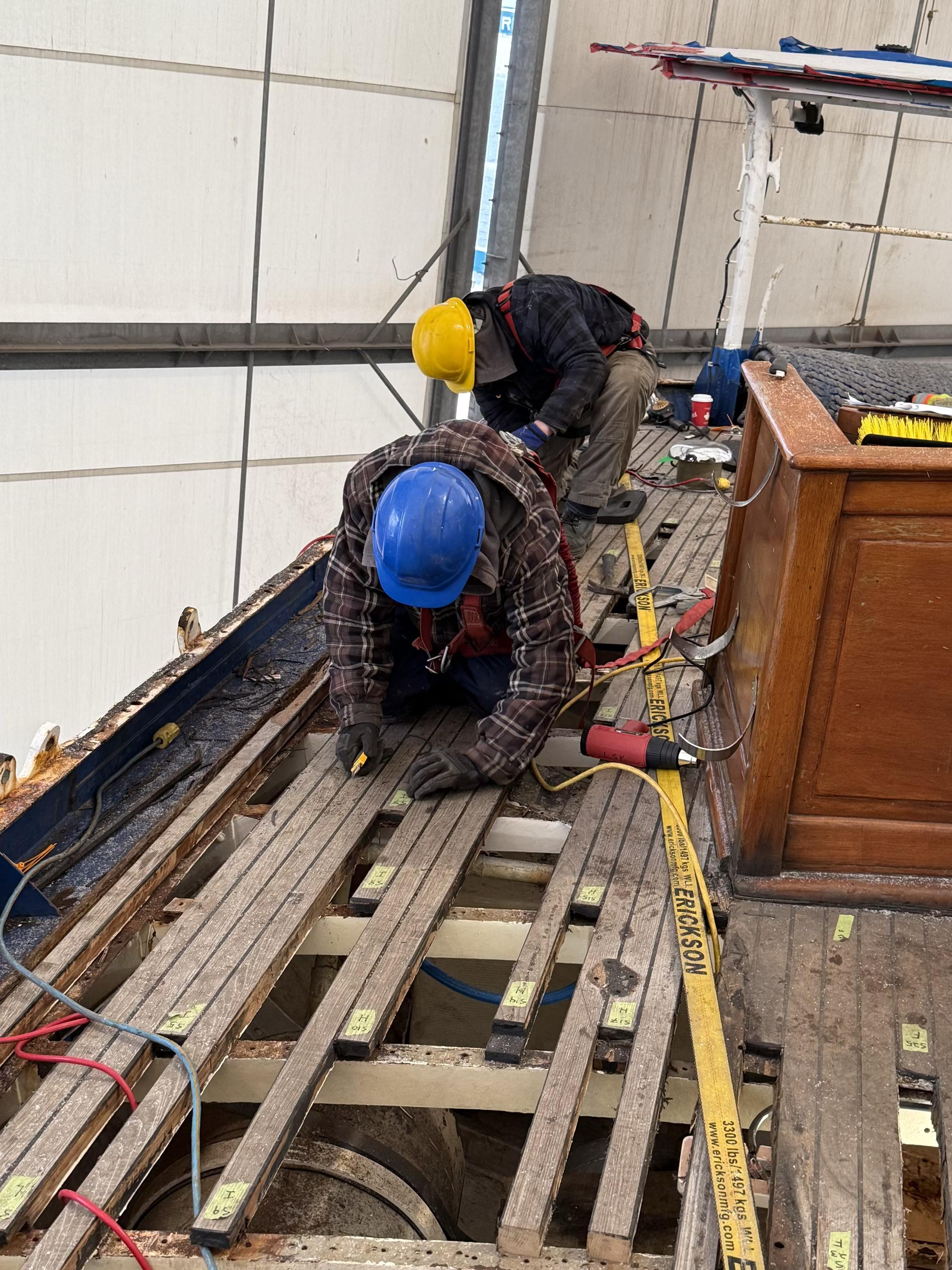 Two construction workers wearing safety helmets, one yellow and one blue, working on a wooden floor frame inside a building. The workers are focused on the task, with tools and yellow caution tape nearby.