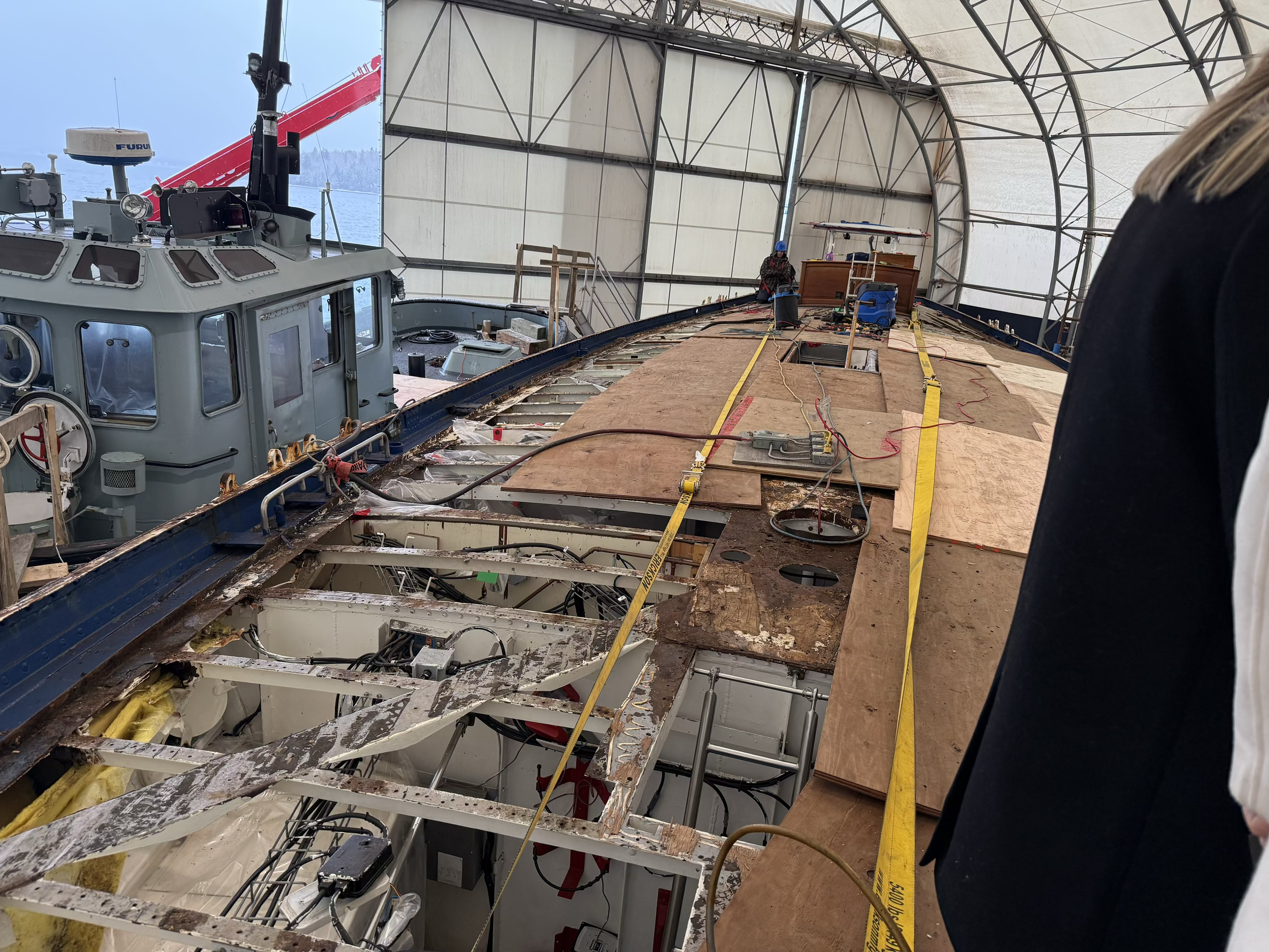 The interior of a boat under maintenance, with exposed wiring, plywood panels, and equipment inside a large shelter. There are workers in the background near the bow of the boat.