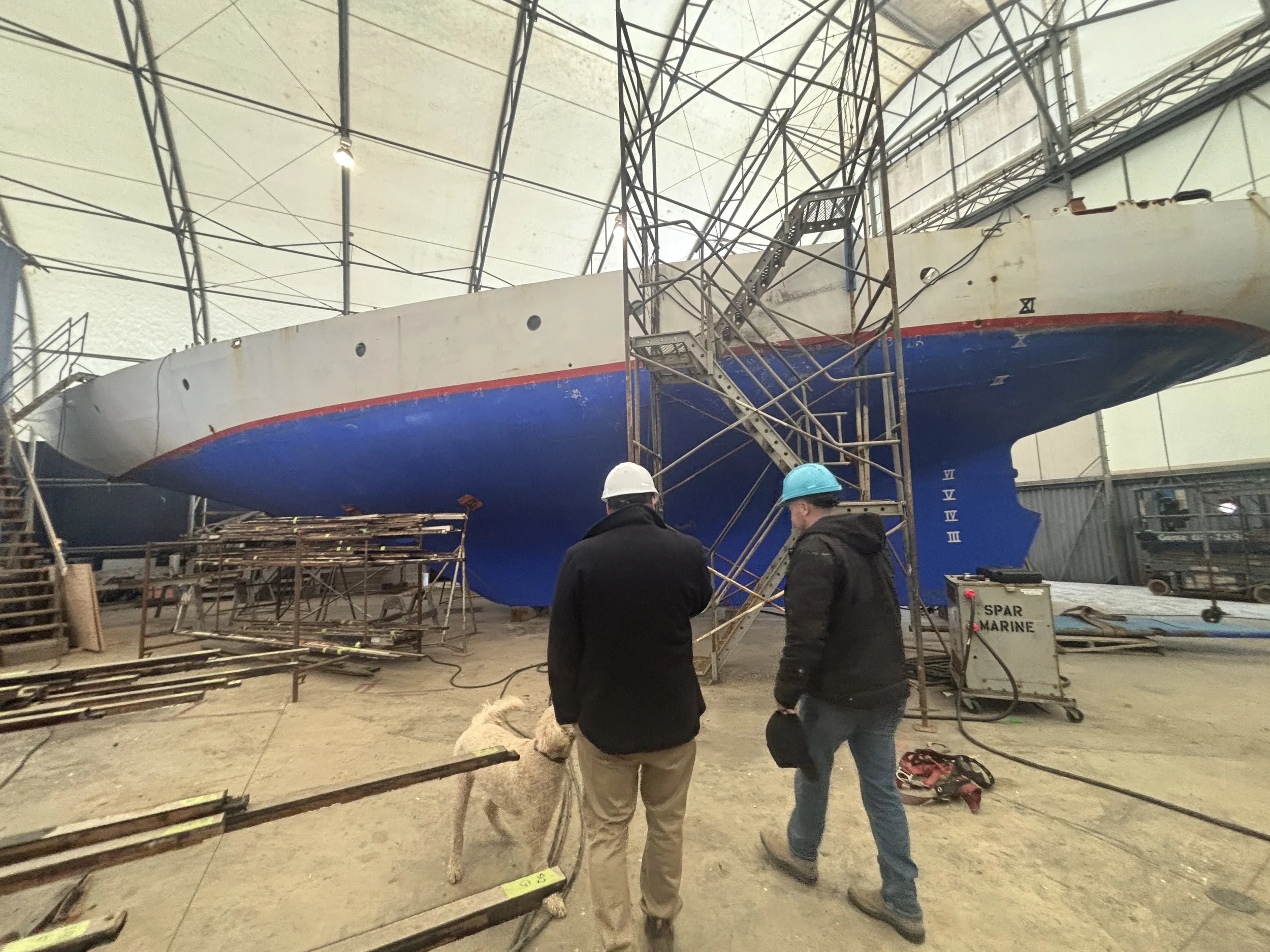 Two workers wearing safety helmets standing near a large ship hull inside an industrial warehouse, with scaffolding and equipment around.