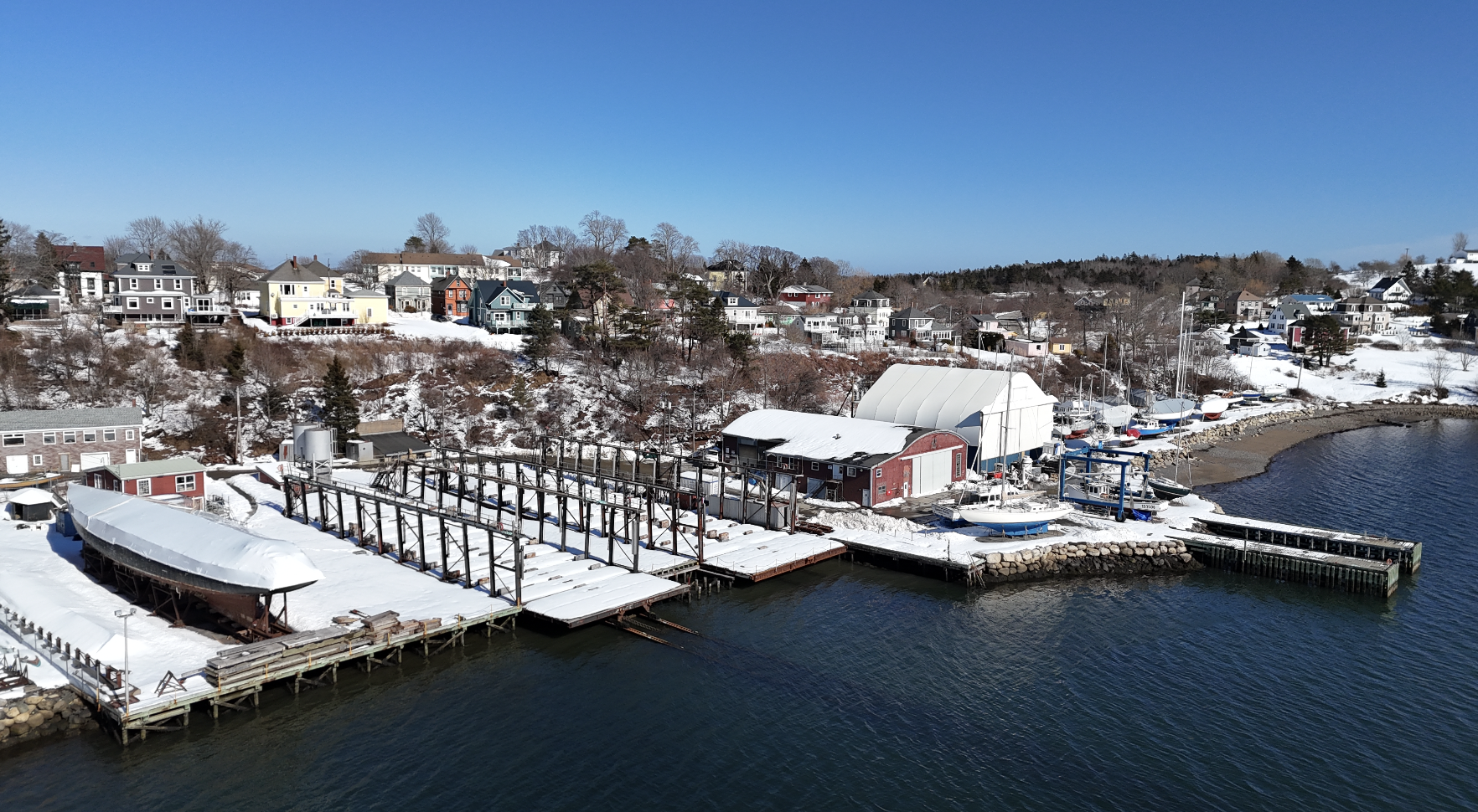 A snowy waterfront scene with boats docked at a marina, surrounded by houses on a hillside under a clear blue sky.