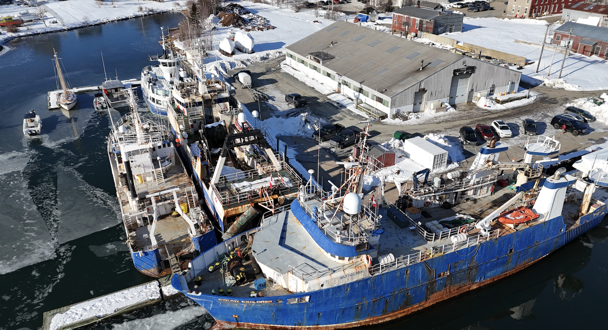 An aerial view of a snowy dock with several boats and ships docked, surrounded by a snowy landscape and parking lot.