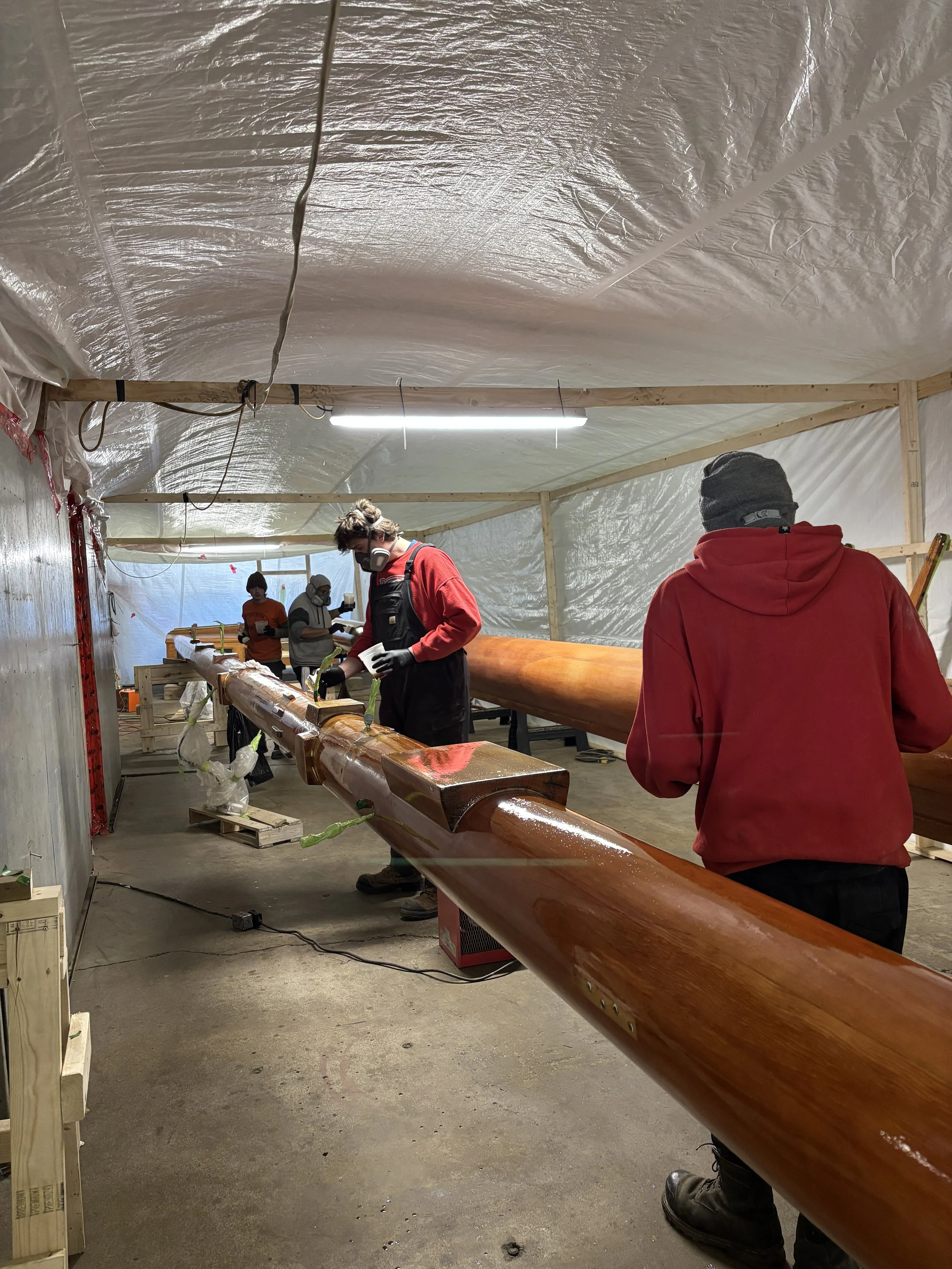 People working on a long wooden kayak inside a workshop, with plastic sheeting on the ceiling and fluorescent lighting.