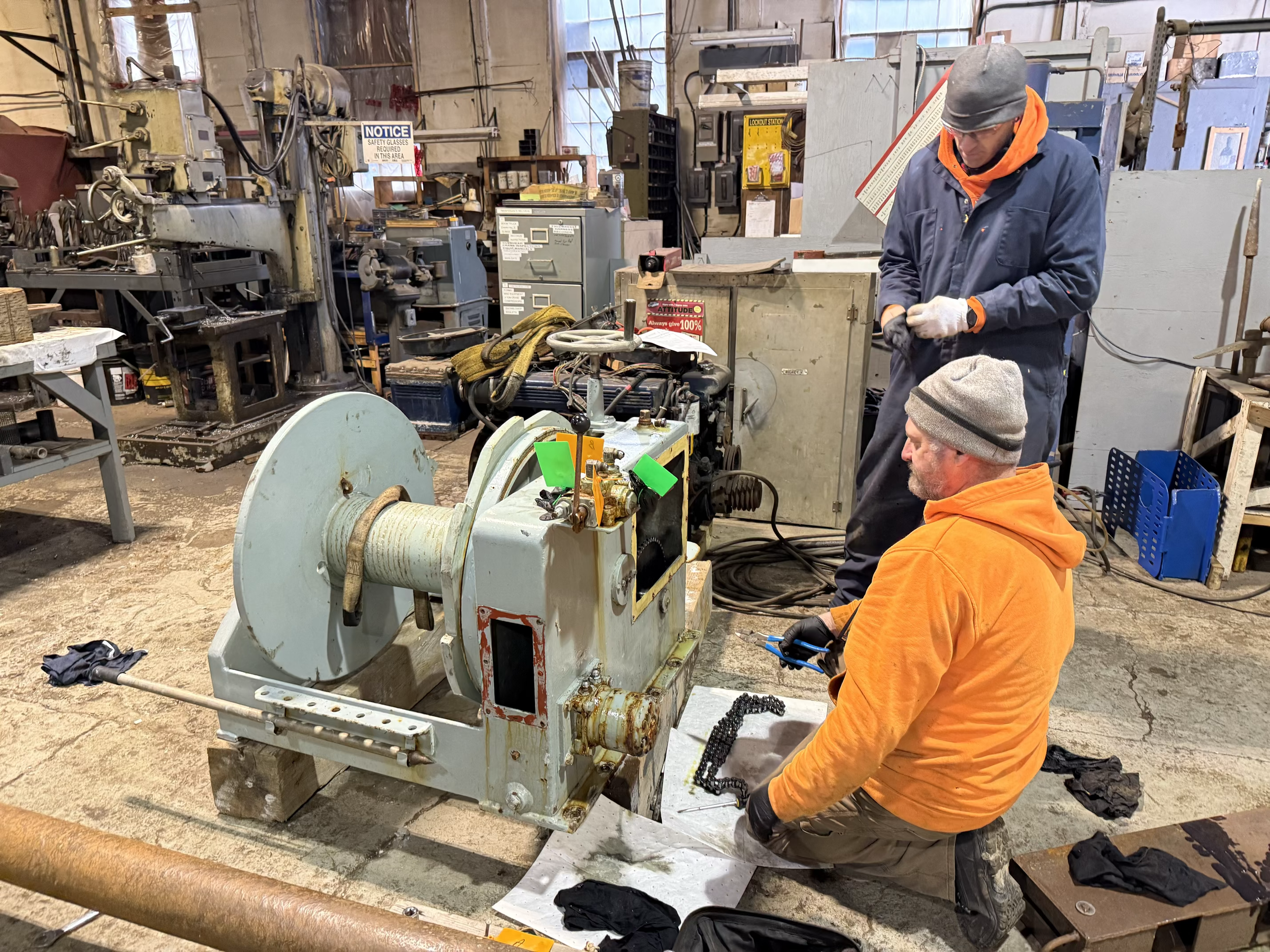 Two men working on a large mechanical device in a workshop. One man is kneeling, wearing an orange hoodie, and the other is standing, wearing a grey beanie and a blue jacket. The workshop contains various tools and machinery.