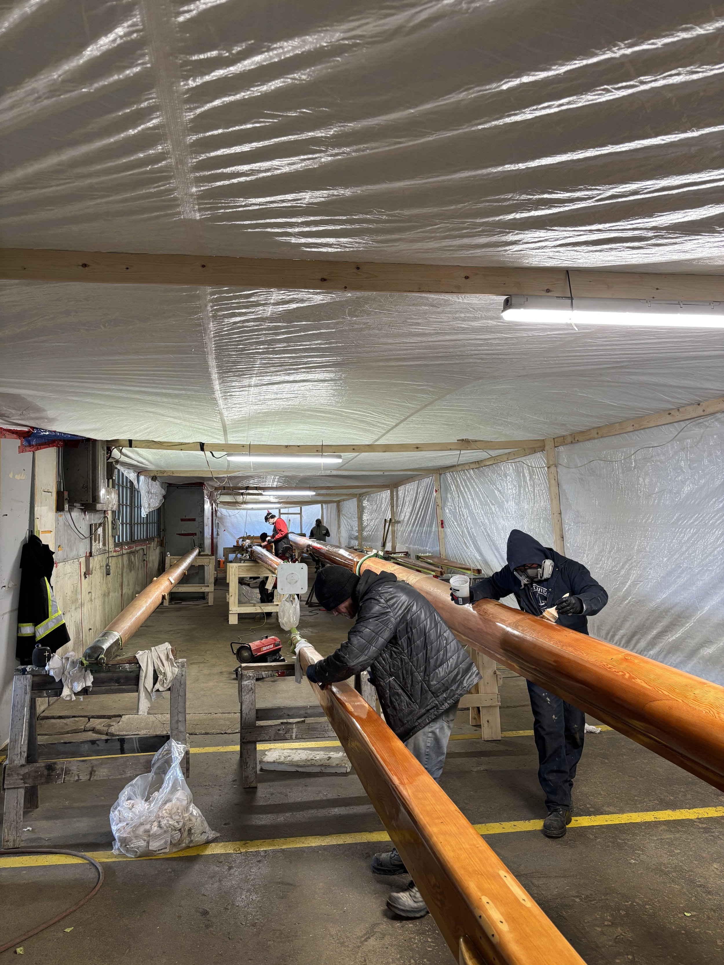 People working on wooden canoe paddles inside a workshop with plastic-covered walls and ceiling.