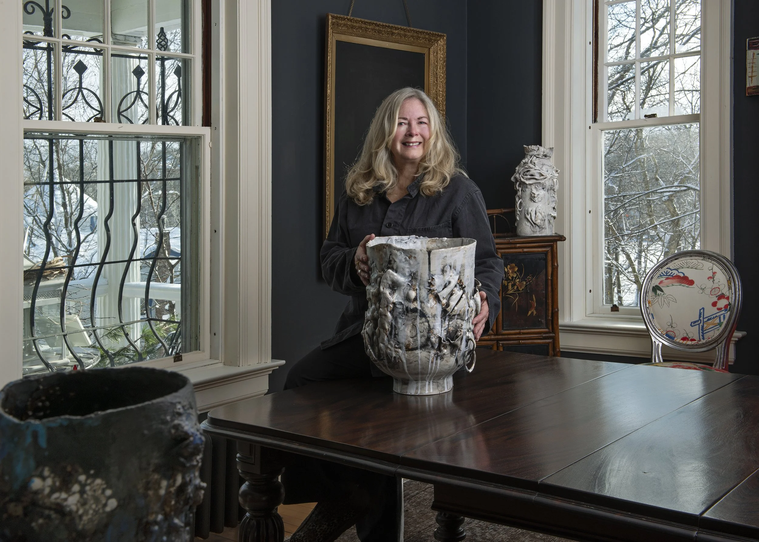 Heather with an artistic ceramic vase in a sunlit room with large windows. The room features dark walls, decorative artwork, and a wooden table.