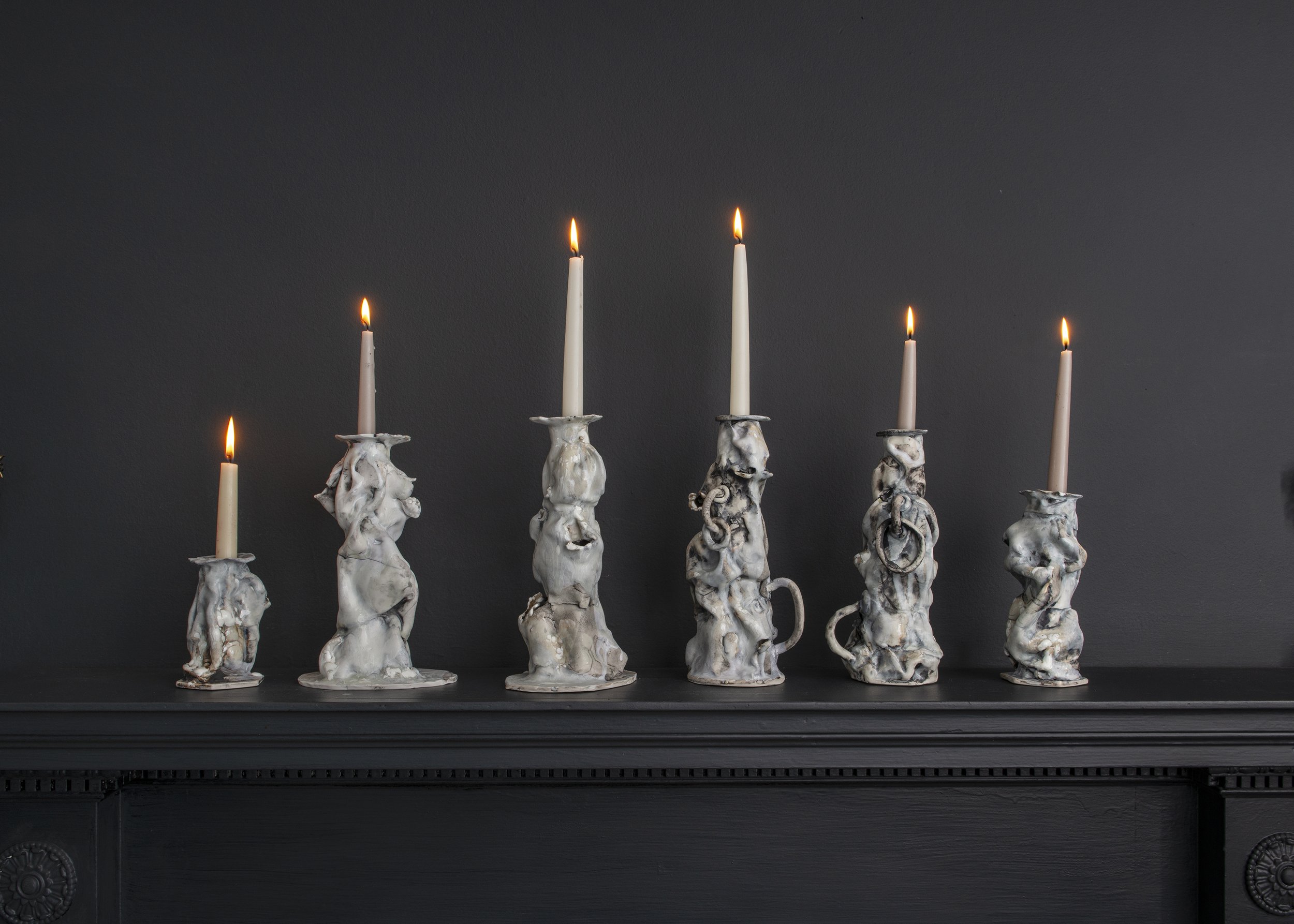 Six white candles with flames on top, held by bone-like candelabras on a black surface against a dark background.